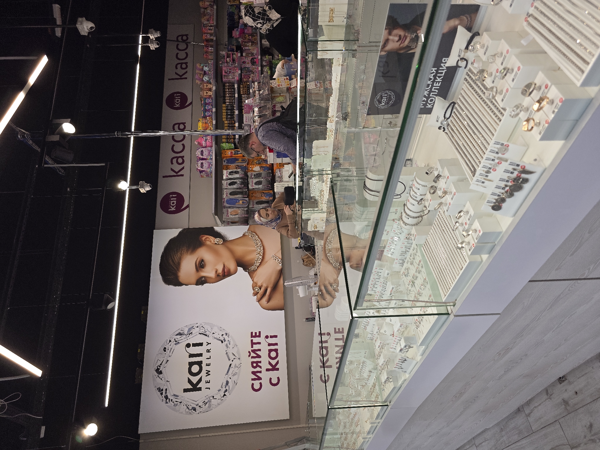 Here is a description of the image:
**Scene Overview:**
The image depicts the interior of a "Kari Jewelry" store, presumably in Grozny, Russia. The store features a display case filled with jewelry, including necklaces, bracelets, rings, and other accessories.
**Key Elements:**
* **Display Case:** The main focus is a well-lit display case showcasing the jewelry. The display is neatly arranged.
* **Advertisements:** A large advertisement is visible on the side of the display featuring a woman wearing jewelry.
* **Store Exterior:** A person can be seen browsing products in the background, which appear to be toys and children's products.
* **Lighting:** The store is well-lit with overhead lighting, including linear LED lights.
**People:**
* **Customer:** A person is seen in the background, likely a customer browsing the store.
* **Advertised Model:** The advertisement features a woman wearing jewelry, likely a model used for promotional purposes.
**Objects:**
* Jewelry, including necklaces, bracelets, rings, and other accessories.
* Toy products
* Display cases
* Advertisements
* Lighting fixtures
**Overall Impression:**
The image captures a typical retail setting with a focus on jewelry sales. The lighting and arrangement of the merchandise are designed to attract customers and showcase the products effectively.