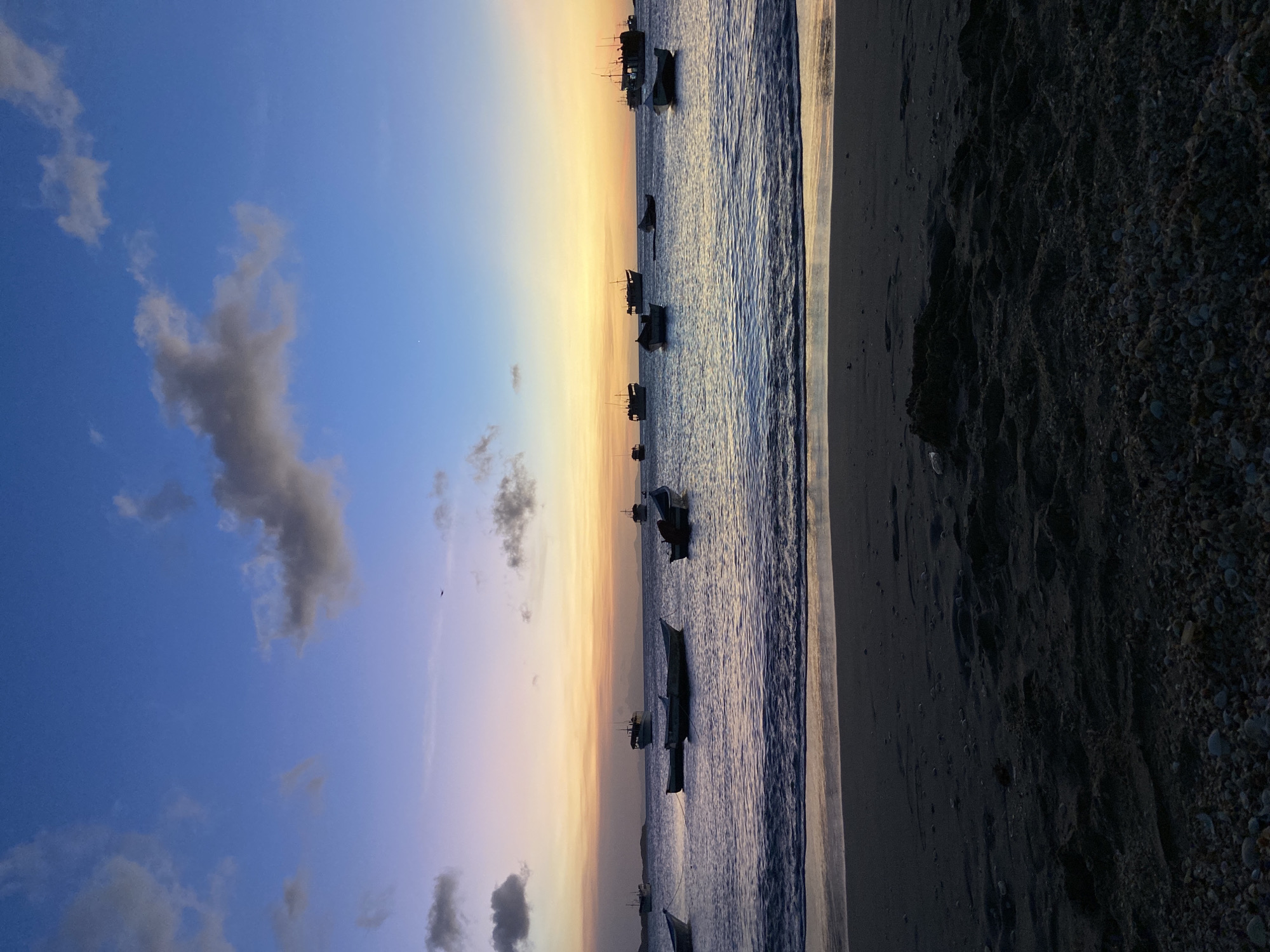 This image depicts a coastal scene in Juan Griego, Venezuela, at dusk. The sky presents a gradient of colors, transitioning from deep blue at the zenith, through shades of purple and pink, to vibrant orange and yellow hues at the horizon. Scattered grey cumulus clouds are visible, primarily in the upper left and right, with smaller wisps across the expanse. A dark, bird-like silhouette is present in the upper middle sky, and a faint white speck, possibly a star or planet, is discernible in the upper right quadrant.
The body of water, likely a bay, is calm with gentle ripples, reflecting the sky's colors, appearing dark blue to purple. Numerous boats are anchored across the water. Approximately five to six smaller, lighter-colored vessels, possibly dinghies or small fishing boats, are positioned closer to the foreground. Further out, against the bright horizon, an estimated eight to ten larger, darker vessels, likely fishing boats or small cargo ships, are silhouetted, some displaying mast structures.
The foreground consists of a dark sand beach interspersed with numerous small shells and pebbles, particularly prominent in the bottom third of the frame. Gentle waves are breaking softly at the water's edge, creating a narrow band of wet sand. No individuals are visible in the scene, and no overt actions or interactions are occurring. Distant, low-lying landmasses are silhouetted against the bright horizon on the far side of the bay.