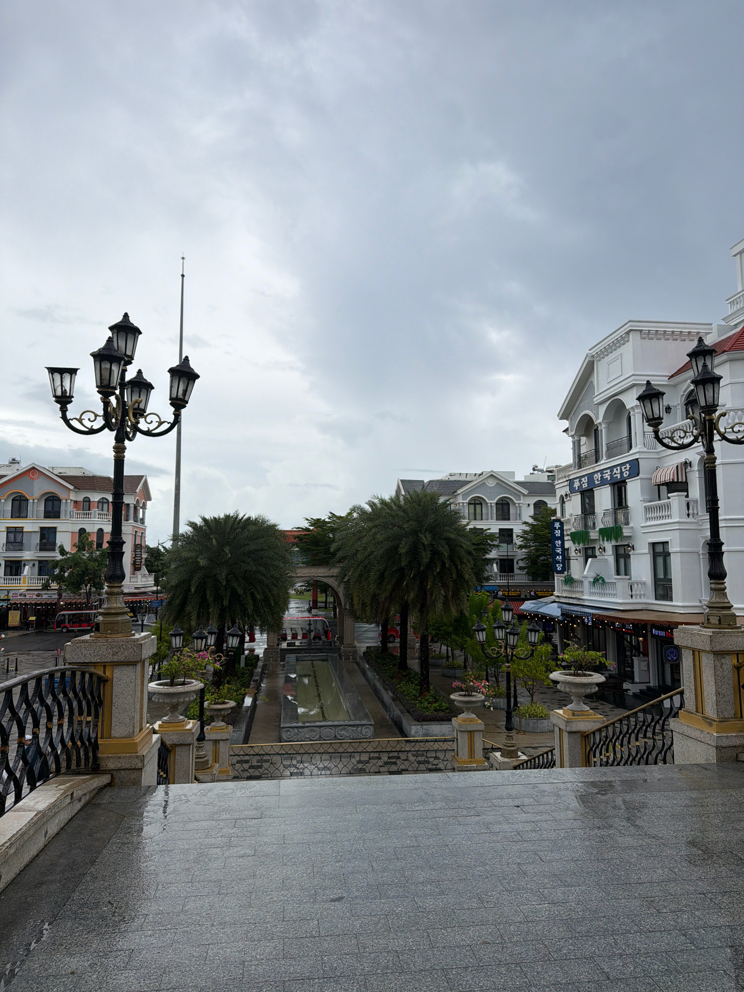 The image depicts a view from an elevated stone staircase looking down onto a street in Phú Quốc, Vietnam. The staircase is wet, suggesting recent rain. On the right side of the frame, a white building with classical architectural elements, including arched windows and balconies, stands prominently. A sign with Korean text, "푸짐 한국식당," which translates to "Pujim Korean Restaurant," is visible on this building. Another building with similar architectural style is seen further down the street. Palm trees line the street, and several vintage-style street lamps are present. The sky is overcast and grey. There are no people clearly visible in the scene. Red tourist trolleys are parked along the street. The overall impression is of a resort town street scene on a cloudy day.