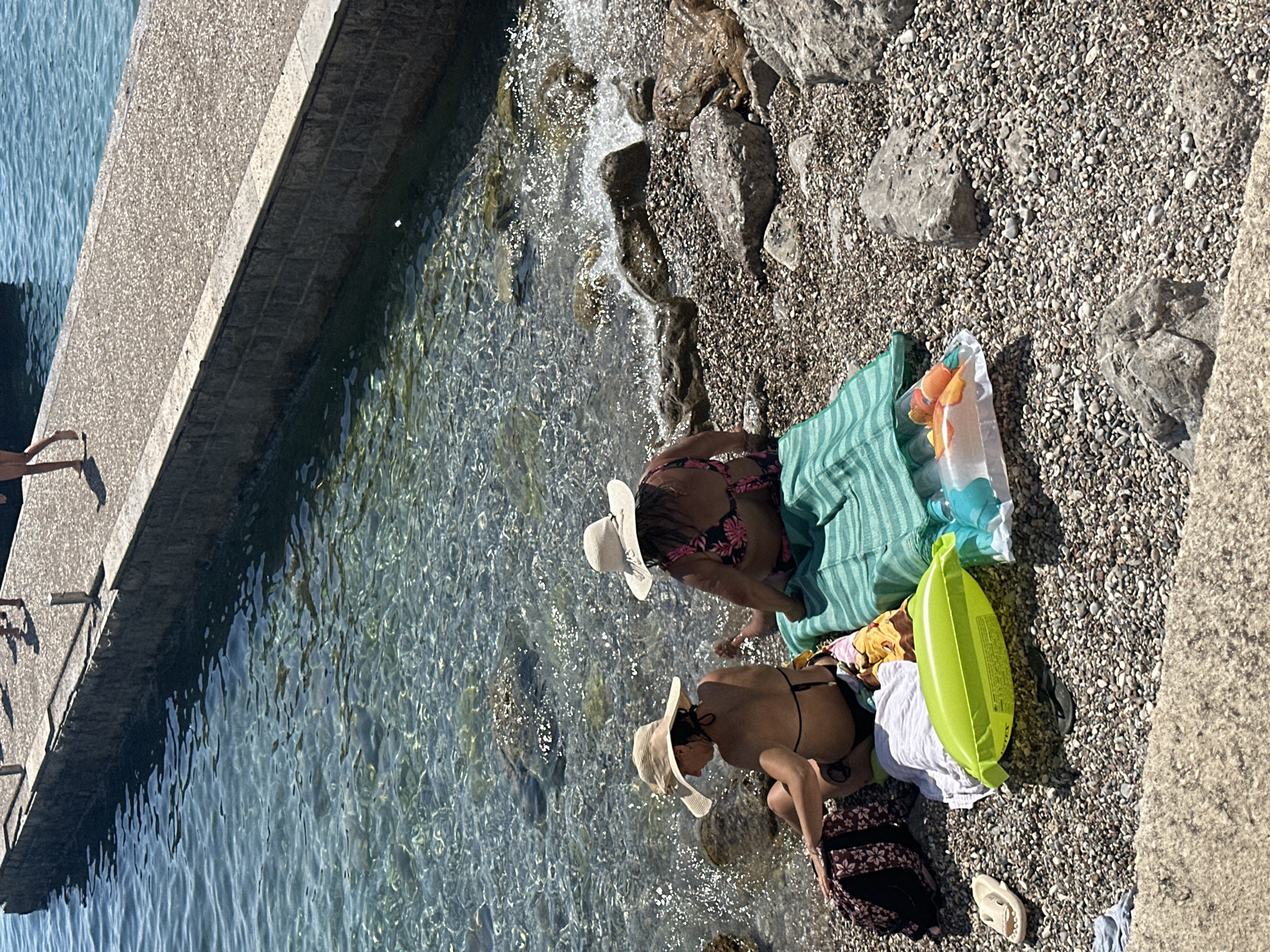 Two women are sitting on a pebble beach in Budva, Montenegro, near the clear turquoise water. The woman on the left is wearing a wide-brimmed straw hat and a black bikini. She is facing left, with her back to the camera. The woman on the right is wearing a white straw hat and a floral bikini. She is facing away from the camera, towards the water. Both women are sitting on towels. In front of them are beach accessories, including a bright green inflatable float, a clear plastic bag with orange objects inside, a white towel, and a patterned bag. The water is shallow and calm, with sunlight reflecting off the surface. A stone wall and a paved walkway are visible in the background, with a few figures walking on the walkway. The beach is covered in small, grey pebbles and larger rocks.