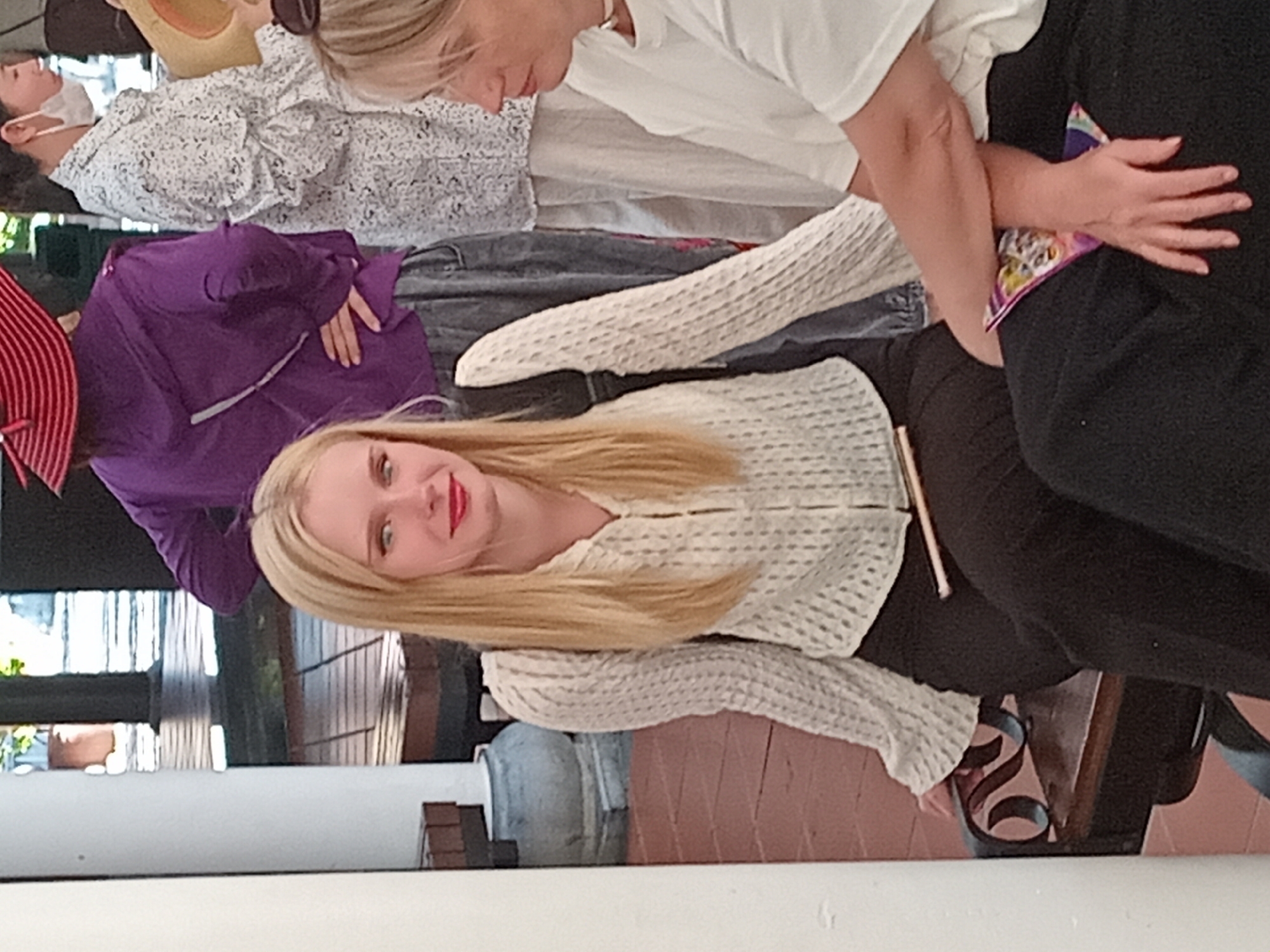 A group of people are gathered outdoors, likely in a public space in Da Nang, Vietnam. In the foreground, a young woman with blonde hair and bright red lipstick is seated on a dark bench, wearing a white, textured long-sleeved shirt and black pants. She is looking forward with a slight smile. To her right, a woman with blonde hair is partially visible, also seated, wearing a white t-shirt. Behind them, a person in a purple jacket and a red and white striped hat stands with their hands on their hips. Another person to the far right is wearing a white patterned shirt and a face mask. The environment appears to be a covered walkway or pavilion, with wooden flooring and decorative pillars.