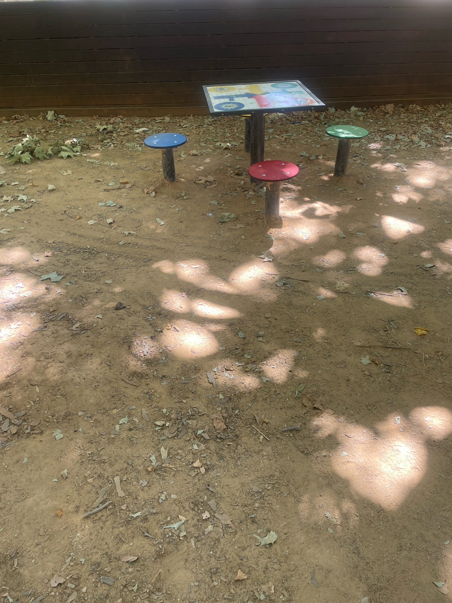 A wooden fence forms the background of an outdoor scene. In front of the fence, a rectangular table with a colorful board game on its surface is supported by two dark, vertical posts. Three round, flat stools surround the table, each mounted on a single post. One stool is blue, another is red, and the third is green. The ground is covered with dirt, scattered leaves, and debris, with dappled sunlight illuminating patches of the surface, suggesting the presence of trees overhead. The overall setting appears to be a park or recreational area. The location is in the city of Atarfe, Spain.