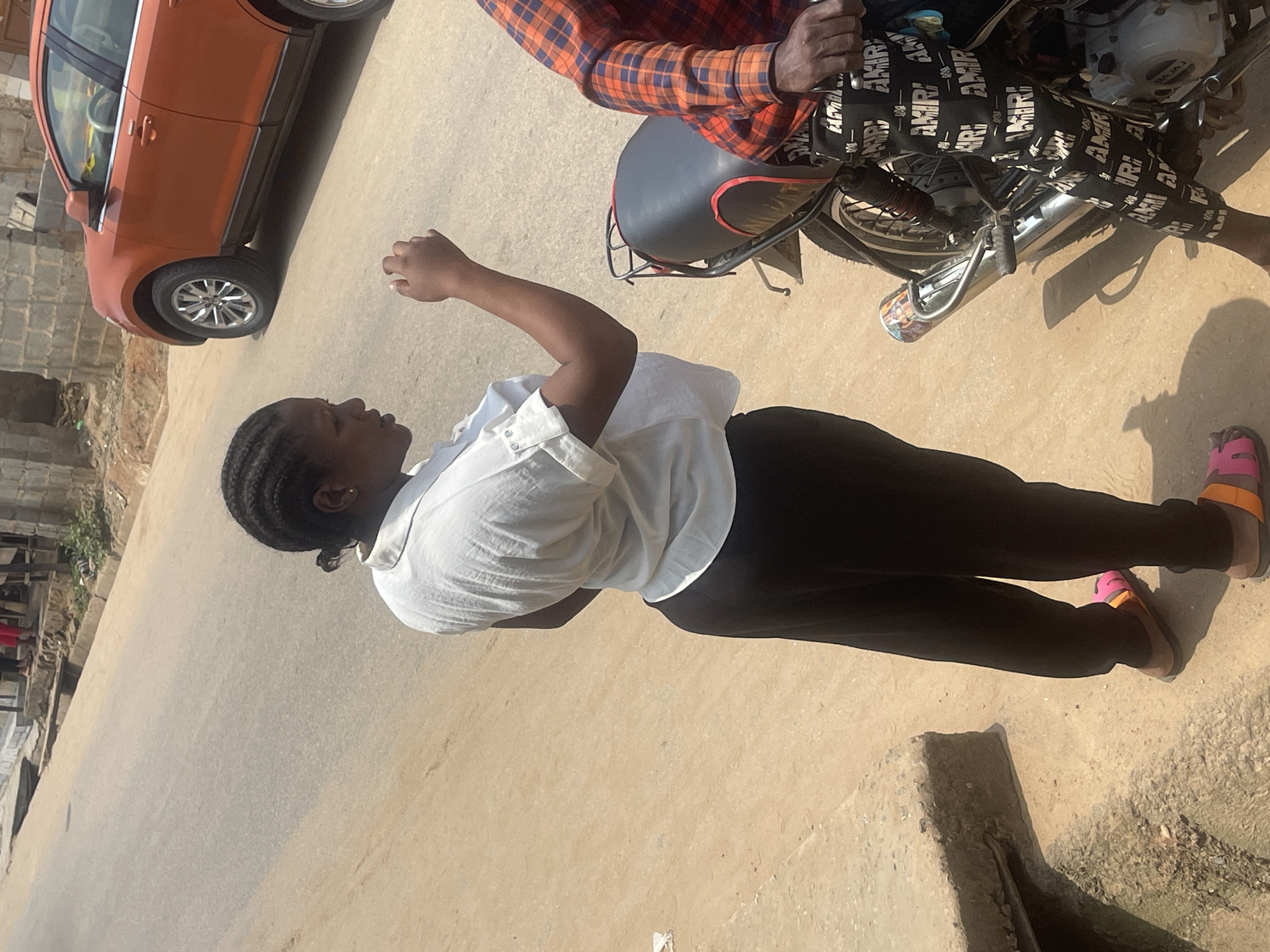 The image depicts an outdoor scene on a paved road in Abuja, Nigeria. A woman with braided hair, wearing a white collared shirt and black pants, stands facing to the right, her left arm raised as if gesturing or speaking. She is wearing pink and orange sandals. Beside her, a person wearing a red and blue plaid shirt and black pants with "AMIRI" printed repeatedly, sits on a motorcycle. The motorcycle has a black seat with red trim, and a silver exhaust pipe. In the background, an orange SUV is parked on the side of the road. The environment appears to be a street with some buildings and vegetation visible. The ground is a mix of sand and asphalt.