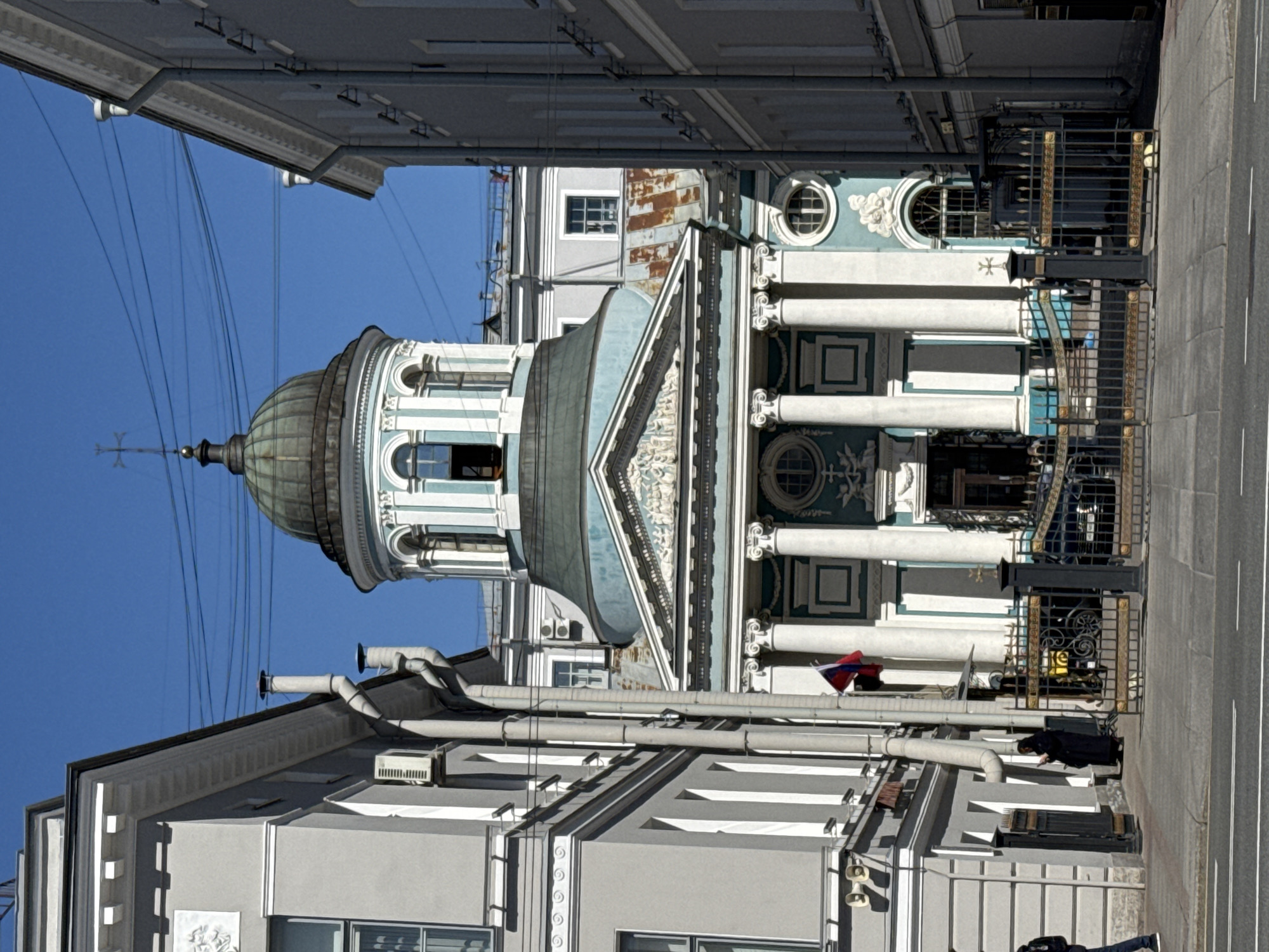 A street-level view shows the St. Catherine Lutheran Church in St. Petersburg, Russia. The church is in the background, featuring a dome, columns, and ornate architectural details. In the foreground, a street is visible. A person is seen near the base of the church; they are facing down and dressed in dark clothing. Building facades frame the church, showing details like windows, gutters, and other architectural elements. Overhead wires stretch across the sky.