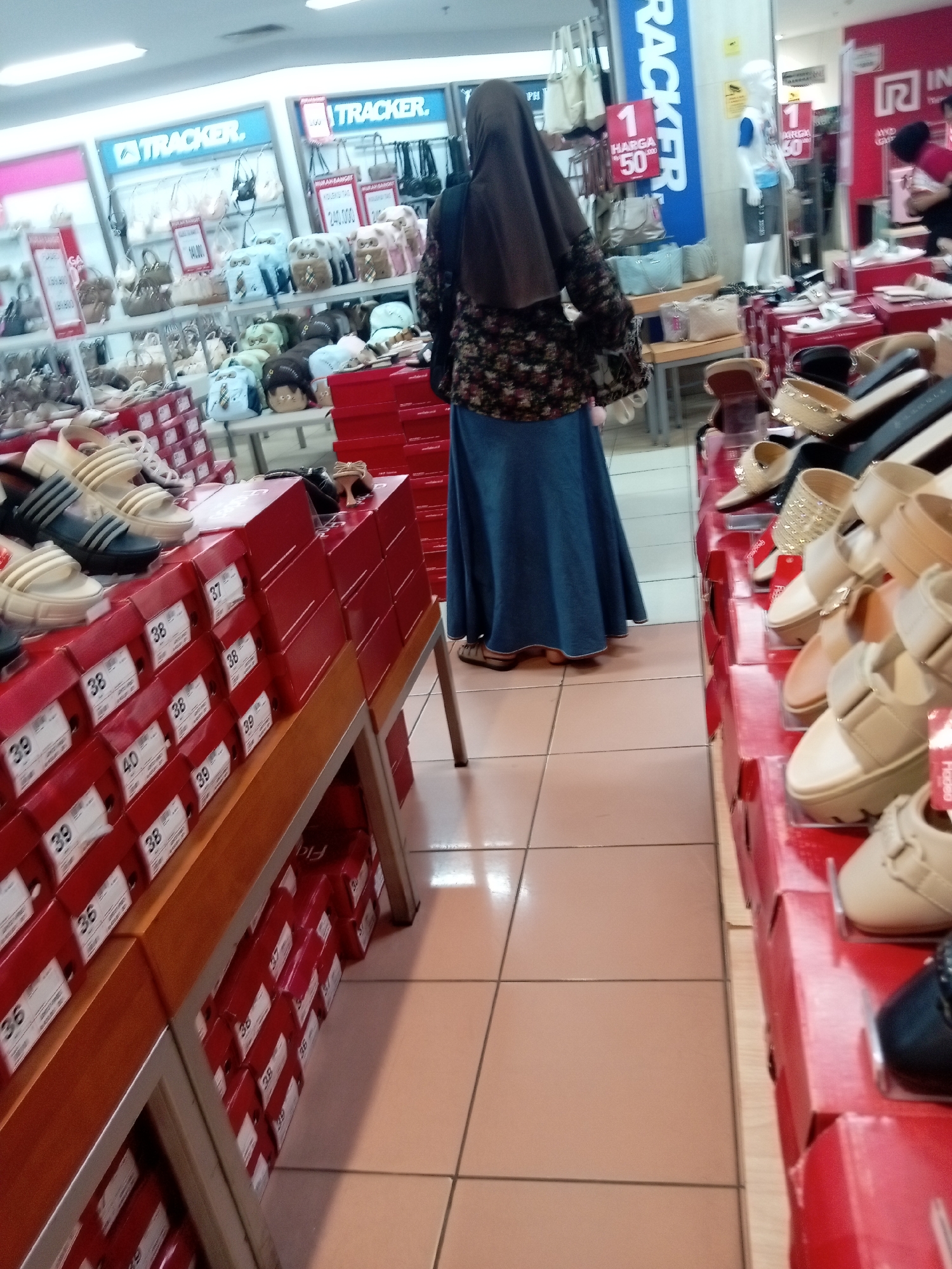 The image shows an indoor retail environment, likely a shoe store, in Batam, Indonesia. In the foreground, rows of red shoe boxes are stacked on wooden shelves. Some boxes display sizes such as "38", "39", and "40". Several pairs of shoes, primarily women's sandals and heels, are displayed on top of these boxes, including beige platform sandals, black heels with embellishments, and cream-colored wedges.
In the mid-ground, a woman wearing a brown hijab and a floral-patterned top with bell sleeves is standing, facing away from the camera. She is wearing a long, denim-blue skirt. Behind her, various handbags and shoes are displayed on shelves, with prominent signage for "TRACKER" visible. Some signs indicate prices like "HARGA 50.000".
Further in the background, a white mannequin wearing a blue shirt and gray shorts stands near a display of more red shoe boxes. Other retail displays with handbags and clothing are visible, and a person in a red shirt is partially visible in the far right background. The flooring appears to be light-colored tiles. The overall impression is of a busy retail setting.