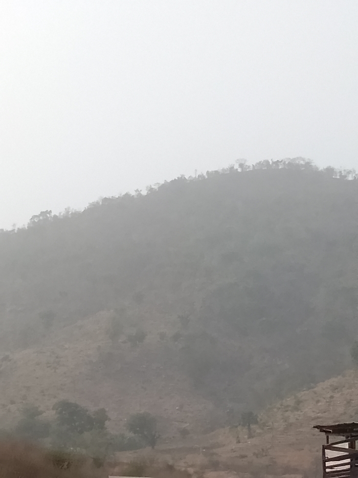 The image depicts a hazy, mountainous landscape in Jibam, Nigeria. A prominent, vegetated hill slope dominates the midground, rising towards a lighter, overcast sky that occupies the upper portion of the frame. The hill's ridge is sparsely dotted with small trees and shrubs, silhouetted against the diffused light. The lower and mid-slopes of the hill are covered in dry, brownish-green vegetation and exposed earth, with further receding hills obscured by atmospheric haze. In the bottom right foreground, a partial view of a rustic, man-made structure is visible, featuring a dark roofline and wooden support beams. No individuals, actions, or interactions are discernible.