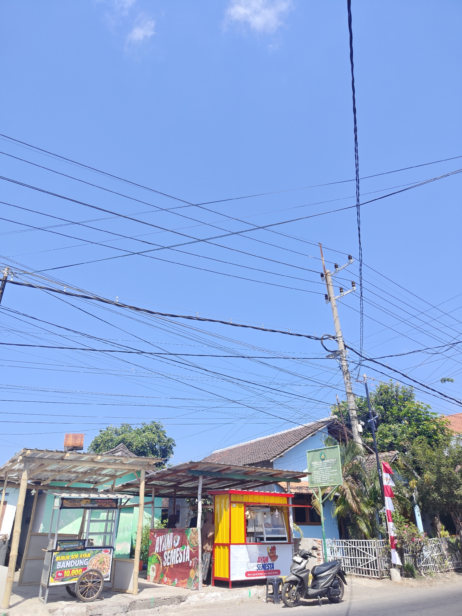 The image depicts a street scene in Situbondo, Indonesia, on a clear, sunny day. A cluster of small food stalls, likely operating from the roadside, are the primary focus. One stall, predominantly green, has a sign advertising "Bubur Sop Ayam Bandung" for Rp. 10,000. Adjacent to it, a brightly colored yellow and red stall features a banner reading "AYAM SEMESTA," with text below indicating "Ayam segra dagan teral panduk terbaik." A motorcycle is parked in front of this stall. Behind the stalls, a blue building with a brown tiled roof is visible, with a green sign displaying text, and a red and white Indonesian flag is mounted on a pole to the right of the building. A network of numerous power lines crisscrosses the clear blue sky, supported by concrete utility poles. Trees and vegetation are present in the background. The scene appears to be a typical urban street environment with small businesses.