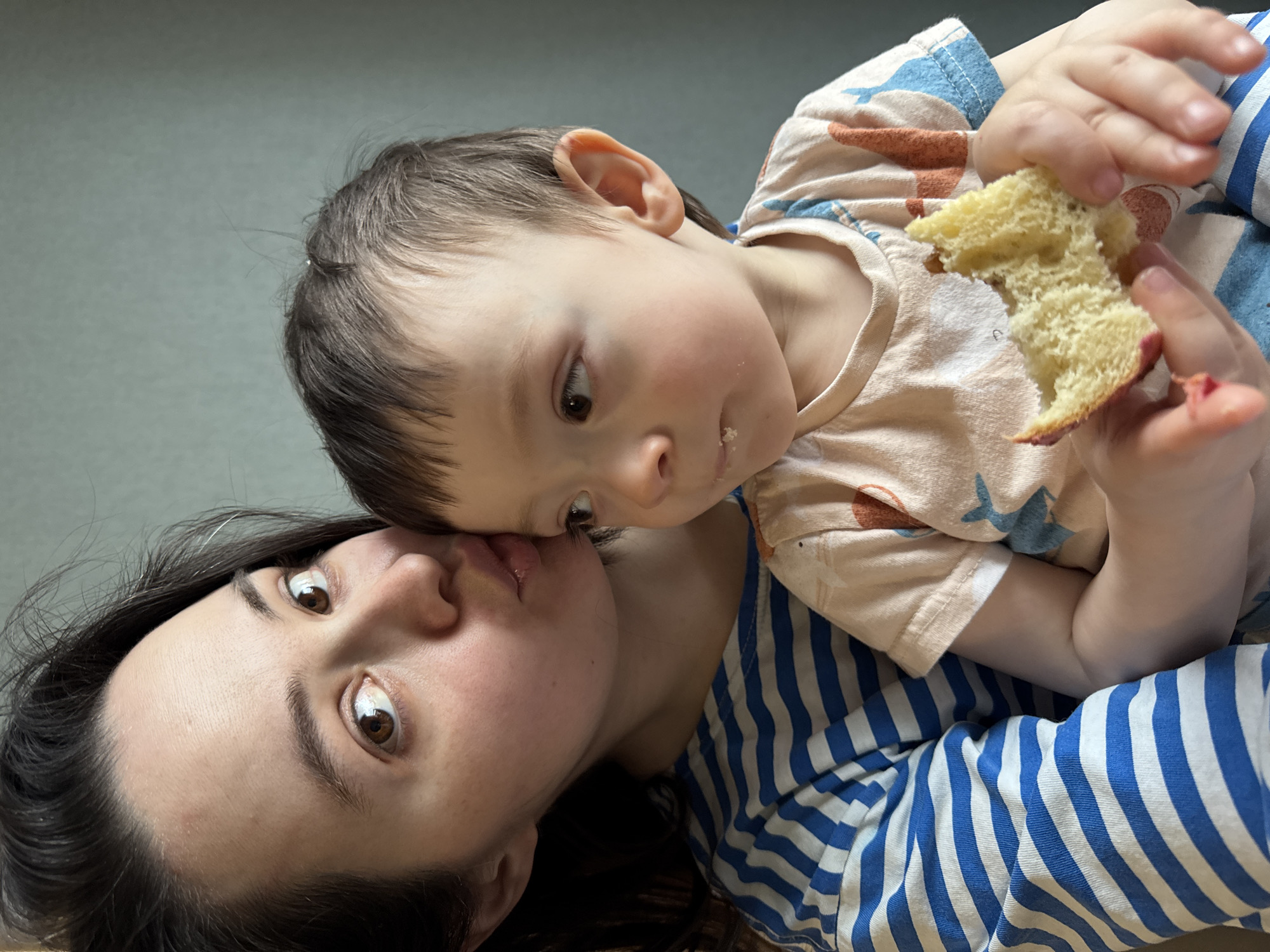 A person with dark hair is lying down, with a child on top. The child is holding a piece of bread. The person and child appear to be indoors. The person is wearing a blue and white striped shirt. The child is wearing a light-colored shirt with brown and blue designs. The location of the photo is unknown, as there are no indicators of the city of Izhevsk, Russia.