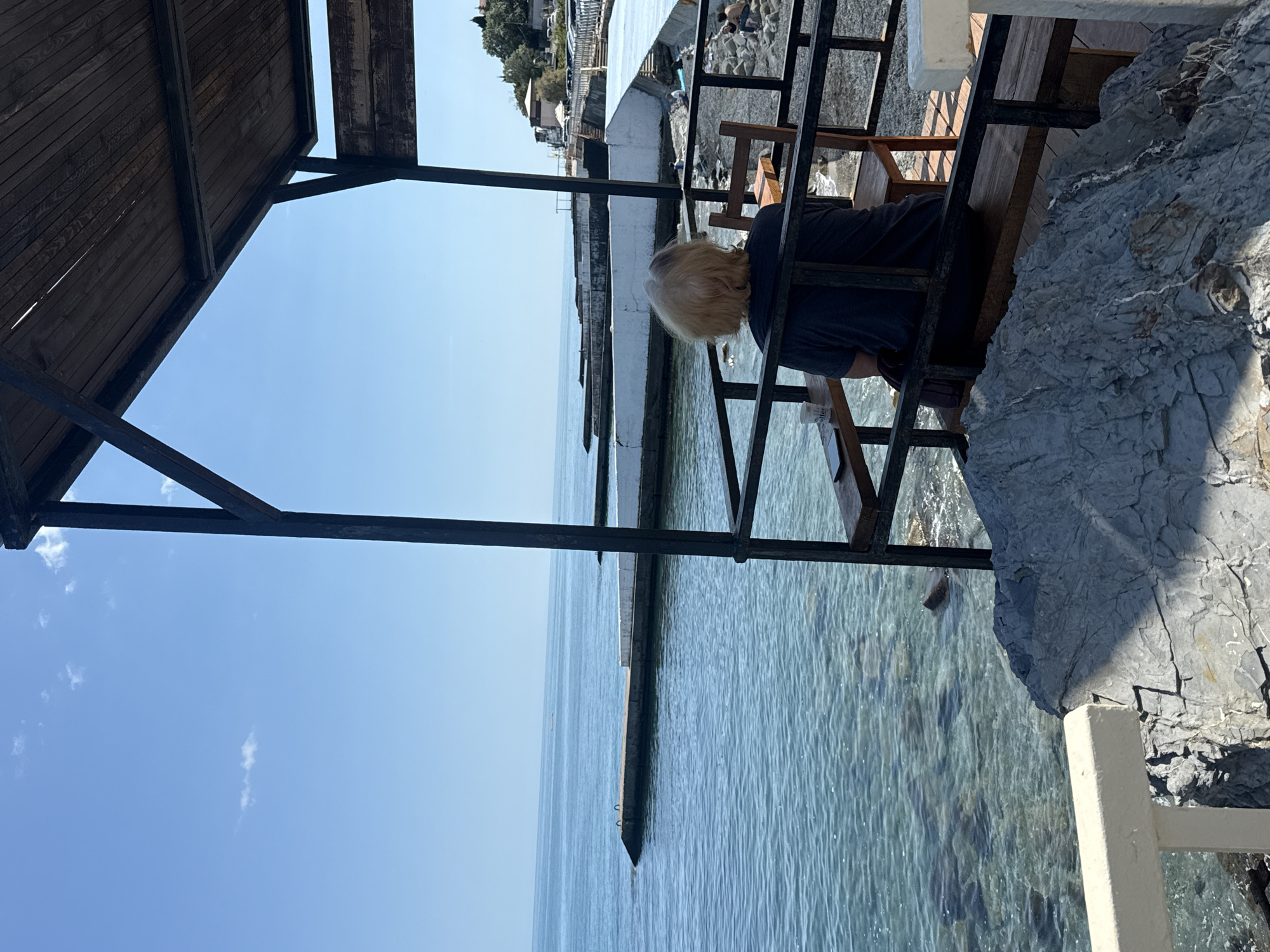 The image captures an outdoor, sunlit scene by the sea, viewed from under a wooden canopy supported by dark metal beams. A person with blonde hair, seen from behind, is seated at a wooden counter or table, facing the calm, blue ocean. They are wearing a dark shirt and appear to be looking down, possibly at a book or device, with their left hand visible. A white disposable cup, faintly bearing the word "coffee," rests on the counter to their left.
To the right of the seated person, a large, textured grey rock formation or cliff face rises from the water. The sea is clear and tranquil, with visible ripples and hints of the seabed in shallower areas. A long, narrow concrete pier or breakwater extends into the distant horizon. Along the mid-ground coastline, several white structures, possibly buildings or parts of a resort, are visible, flanked by green vegetation. Further back, a road with parked cars and more buildings can be discerned. The sky overhead is a bright, clear blue with a few scattered, wispy white clouds, indicating a bright, sunny day, likely midday or early afternoon. The overall setting suggests a peaceful coastal location, possibly a cafe or viewing point overlooking the water.
