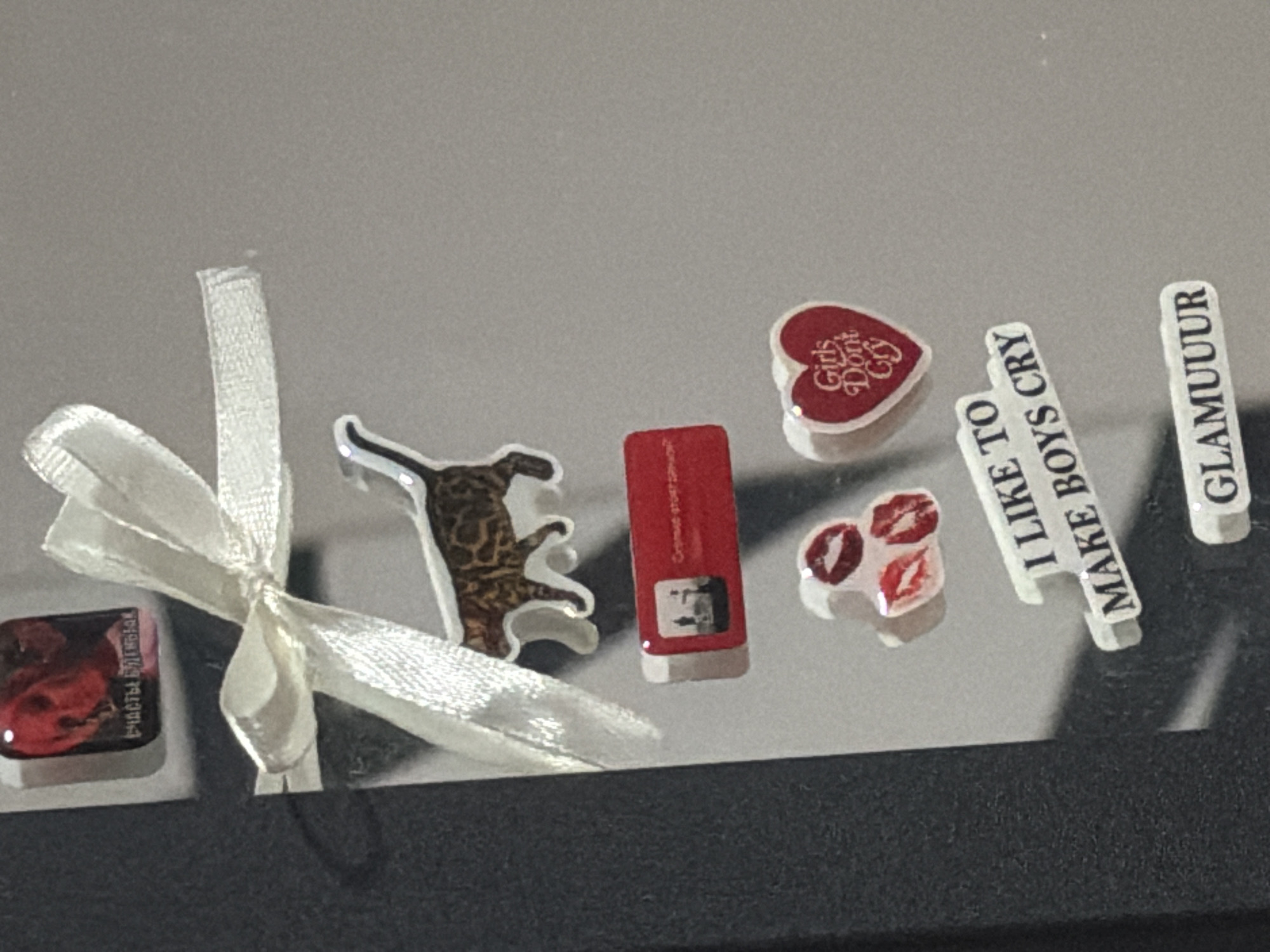 The image displays a collection of stickers and magnets affixed to a dark, textured surface, possibly a refrigerator or a locker. A white satin ribbon is tied in a bow, partially obscuring a magnet depicting a Bengal cat. Below the cat magnet, a rectangular red magnet features a miniature image of a cityscape with two figures standing, and text that appears to be in Cyrillic. Further down, a cluster of three red lip-print stickers is visible. Below these, a white sticker with blue lettering reads "I LIKE TO MAKE BOYS CRY". Another white sticker, positioned lower, displays the word "GLAMUUUR" in black lettering. At the top left, a square magnet shows a red image with Cyrillic text that translates to "HAPPINESS IS CLOSE". The overall scene is static and appears to be a personal decoration. There is no discernible location in Moscow, Russia mentioned or indicated in the image.