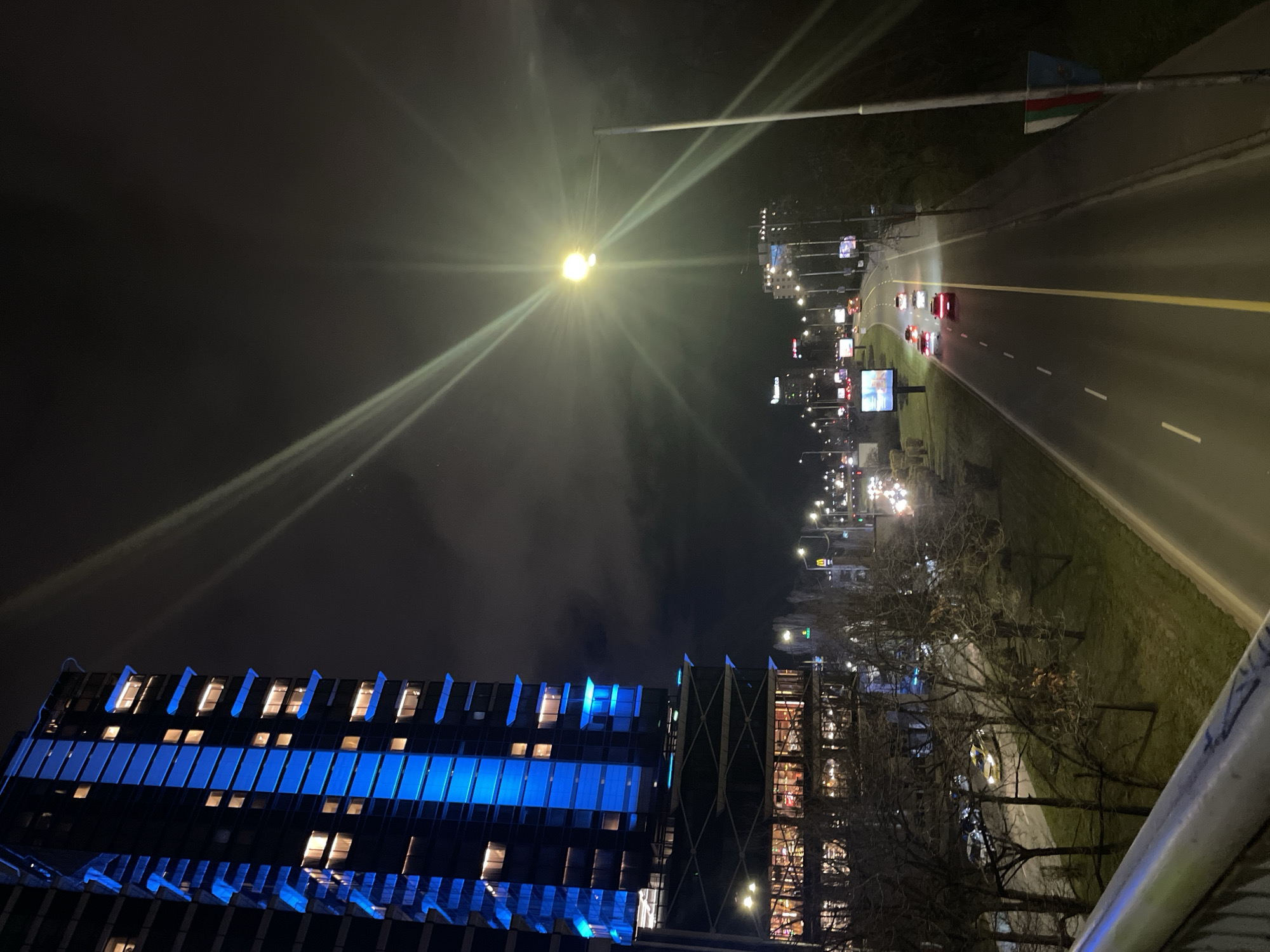 A nighttime cityscape in Sofia, Bulgaria, shows a modern building illuminated with blue lights, and a highway with moving traffic. The highway is brightly lit by streetlights and lined with billboards. A sidewalk and trees run alongside the highway. A light pole with an attached flag stands over the highway. The image appears to have been taken from an elevated position, possibly a bridge. The sky is dark and cloudy.