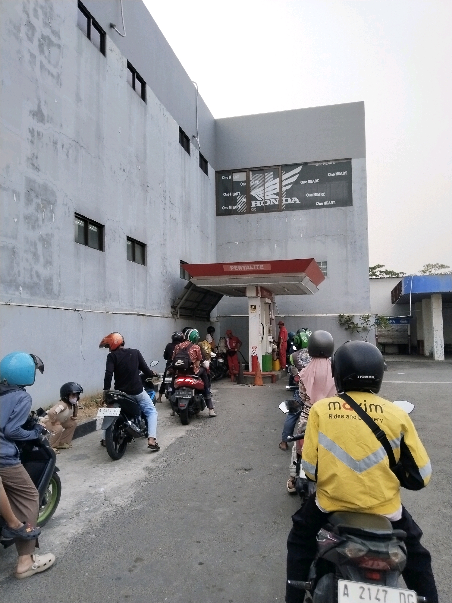 The image depicts a scene at a Pertamina gas station in Serang, Indonesia, where several people on motorcycles are queuing to refuel. The gas station has a red awning with "PERTALITE" written on it. A Honda advertisement is visible on the side of the building. The overall environment is an outdoor urban setting with a building and a paved road.