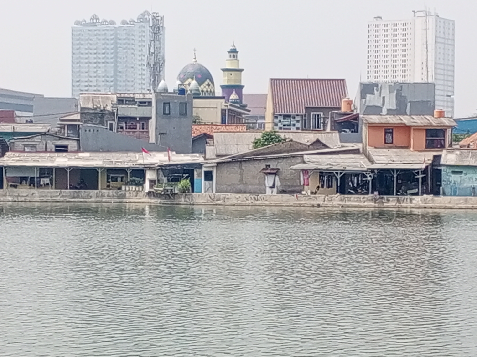 The image depicts a waterside scene in Depok, Indonesia, showcasing a mix of older, low-lying structures and modern high-rise buildings. In the foreground, a body of water with ripples stretches across the frame. Along the embankment, a line of modest houses and shops, some with corrugated metal roofs and open fronts, are visible. A mosque with a dome and minaret stands out amongst these buildings. In the background, two tall, modern apartment buildings dominate the skyline, one on the left with a distinctive tiered top, and another on the right, a stark white rectangular structure. The overall impression is one of urban development, with older residential areas situated near contemporary high-rise constructions in Depok.