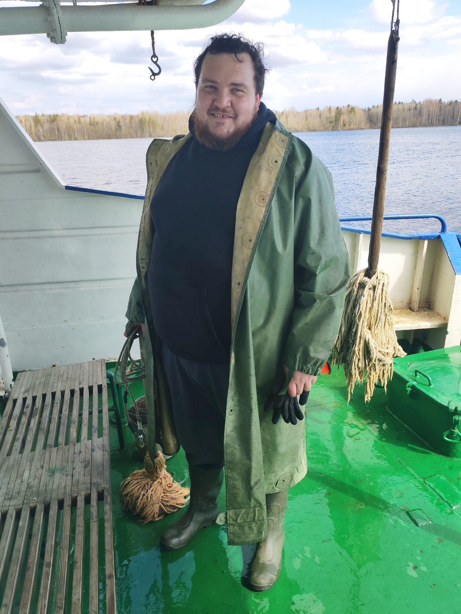 A man stands on the green deck of a boat, posing for a photograph. He is a Caucasian male with a beard and dark, curly hair, smiling pleasantly at the camera. He wears a dark hoodie, dark trousers, a long, olive-green waterproof raincoat with a lighter-colored lining, and green rubber boots. In his left hand, he holds a pair of black work gloves, and in his right, he carries a coil of rope.
The boat's deck is a bright green, appearing wet in some areas. To the left, there's a wooden grate on the deck. To the man's right, a wooden pole with a rope mop head is visible, alongside what appears to be a bucket. The boat has white railings with blue trim, and overhead, a white pipe and a rusty metal hook are suspended.
In the background, a wide body of water stretches out under a cloudy sky with hints of blue. A distant shoreline is lined with trees, many of which appear bare or with very sparse foliage, suggesting an early spring setting. The overall scene depicts a daytime shot on a body of water, likely a lake or wide river, with the man appearing content in his working environment. There is no visible text in the image.