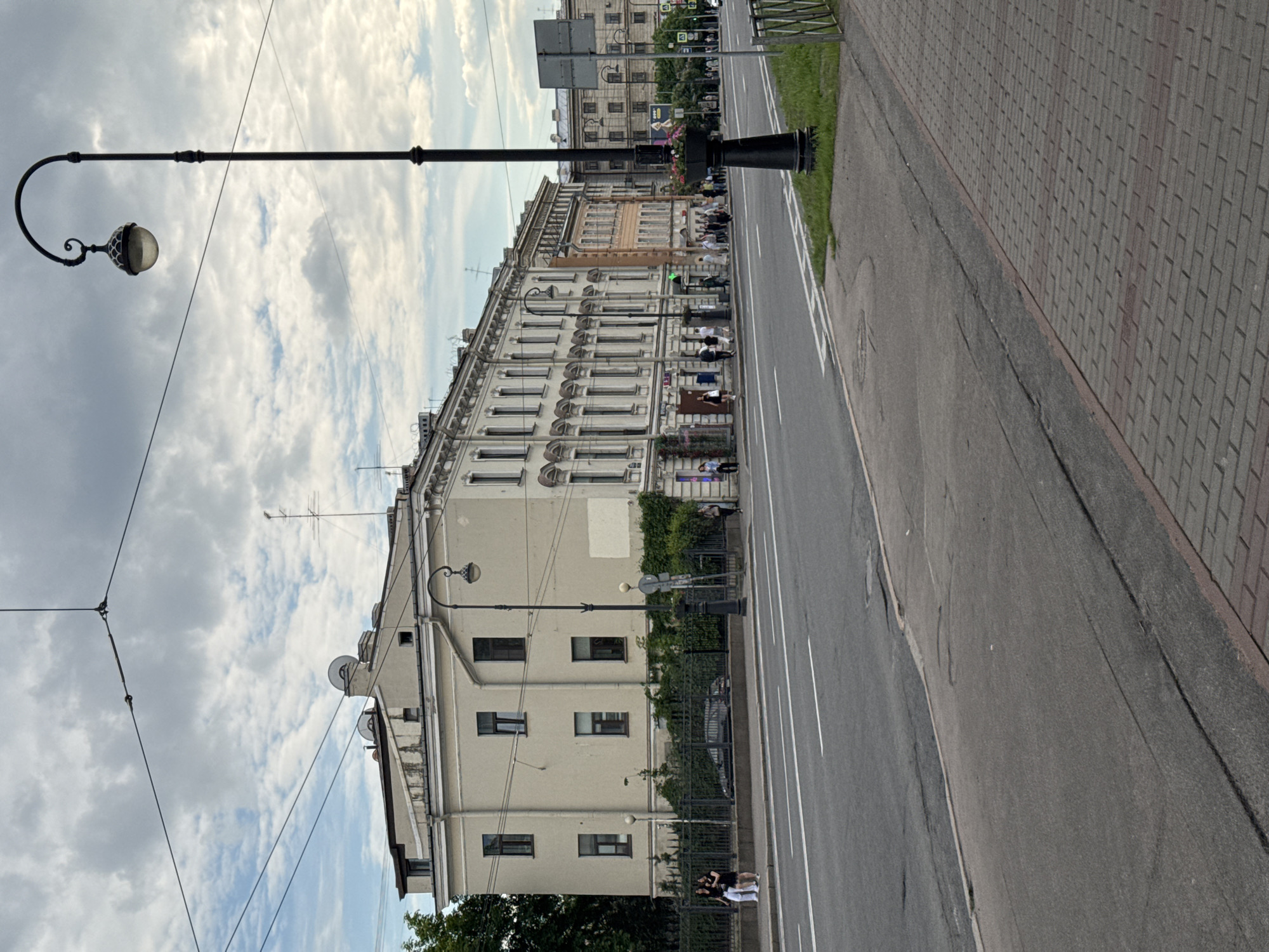 The image captures a vibrant street scene in Saint Petersburg, Russia, under a partly cloudy daytime sky. The perspective looks along a wide street, with a grand, multi-story building dominating the left side of the frame and a road and paved sidewalk on the right.
The prominent building features a light-colored facade with numerous windows, some adorned with decorative elements or small balconies. Along its ground floor, several outdoor dining or cafe areas are visible, with groups of people seated at tables, enjoying the pleasant weather. Further along the building, green climbing ivy covers parts of the wall and a fence. Satellite dishes are noticeable on the upper parts of the building, suggesting residential or mixed-use occupancy.
In the foreground on the left, a tall, black lamppost with an ornate, curved arm holding a spherical light fixture stands out against the sky. The asphalt road in the middle features clear white lane markings, indicating traffic flow. A narrow grassy median strip separates the road from a distant, less distinct building further down. The right side of the image shows a grey brick-patterned sidewalk.
Several individuals and small groups of people are present throughout the scene. Besides those seated at the outdoor cafes, two people are seen standing or sitting near a fence on the left, and others are walking or standing further down the street. No discernible text is visible on signs or buildings due to the distance. The overall impression is one of a typical urban day, with people engaging in leisure and daily activities in a historically rich architectural setting.