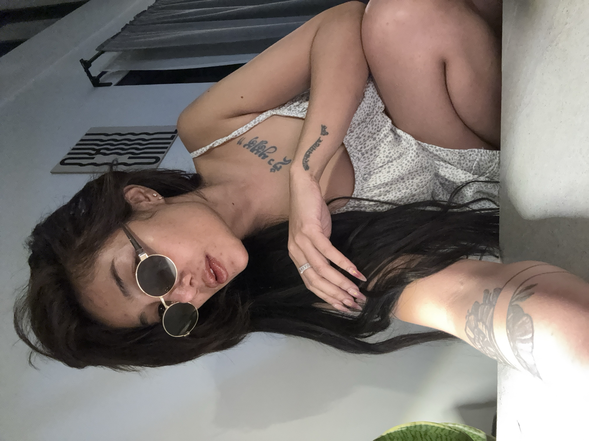 A woman is depicted reclining indoors, captured in a full-body shot rotated 90 degrees clockwise. She is positioned on her side, with her head towards the left of the frame and her body curled inwards. She wears a light-colored, spotted or patterned dress with thin straps. Her long, dark hair cascades around her face and shoulder.
She wears dark, round sunglasses with thin metal frames, partially obscuring her eyes. Her lips are slightly parted. A small stud earring is visible in her left earlobe, and a ring with multiple small stones is on a finger of her left hand.
Several tattoos are visible: one on her left rib cage appears to be in a script, likely Sanskrit or Hindi, resembling "ॐ भूर्भुवः स्वः". Another tattoo, also in script, is on her left forearm, possibly reading "रामनवमी". A larger, dark tattoo, possibly depicting wings or a detailed motif, extends around her upper right arm or shoulder.
The background includes a light-colored wall, with a piece of rectangular wall art featuring black wavy lines on a white background. Above this, horizontal blinds or slats are visible. A partial view of a green plant, possibly a snake plant, can be seen in the bottom right corner of the image. A plain white surface occupies the right side of the frame, possibly a bed or another wall. The lighting suggests an indoor setting, likely daytime or well-lit. The woman is posing, looking towards the camera with a neutral to slightly contemplative expression.