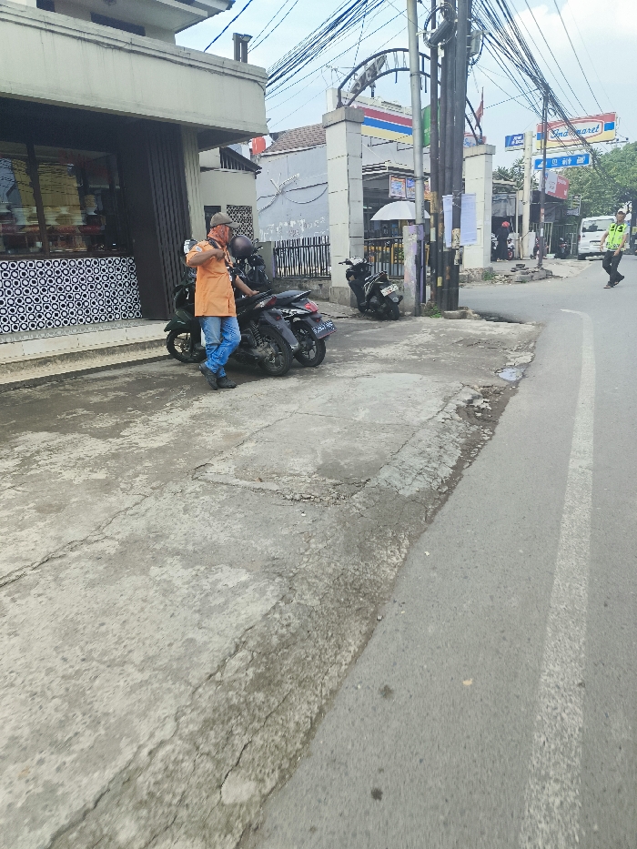 A man wearing an orange shirt and a black face mask stands next to a black motorcycle parked on a concrete sidewalk. He is facing away from the camera and appears to be looking at the motorcycle. To the left of the man is a black motorcycle with a white sticker on the back. The man is standing near a small building with a large window displaying a black and white pattern. There is a single white painted lane on the road. The image was taken near a street intersection in Padalarang, Indonesia. The street is paved with asphalt and the sidewalks are cracked concrete. There are utility poles on the sidewalk. The image is taken from a low angle. The day is overcast.