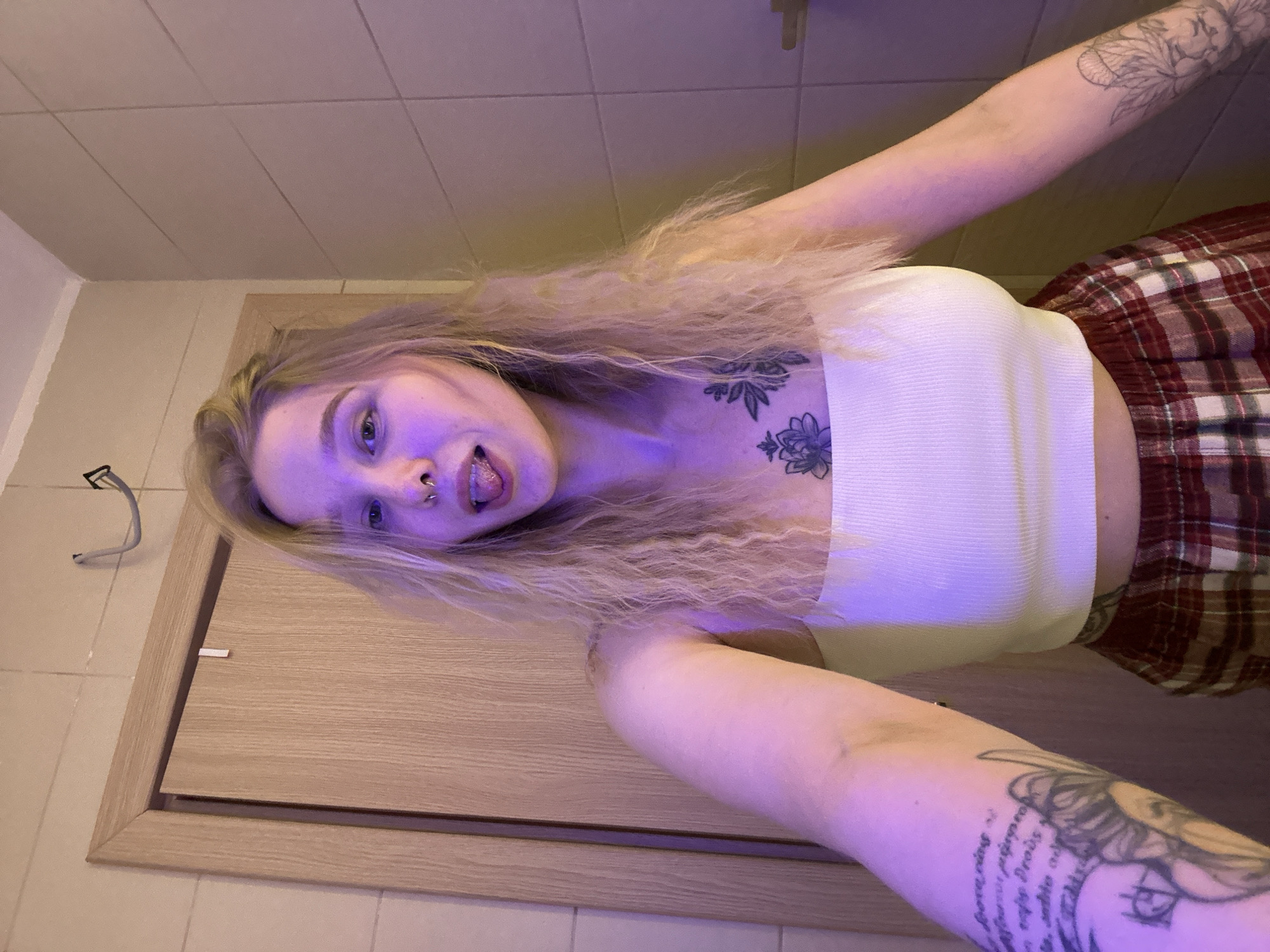 The image shows a woman in what appears to be a bathroom. She has light blonde hair, fair skin, and visible tattoos on her arms and chest. She is wearing a white tank top and a red plaid skirt or shorts. She is sticking her tongue out. The background consists of a beige door and white tiled walls. The location in Budva, Montenegro is unknown.