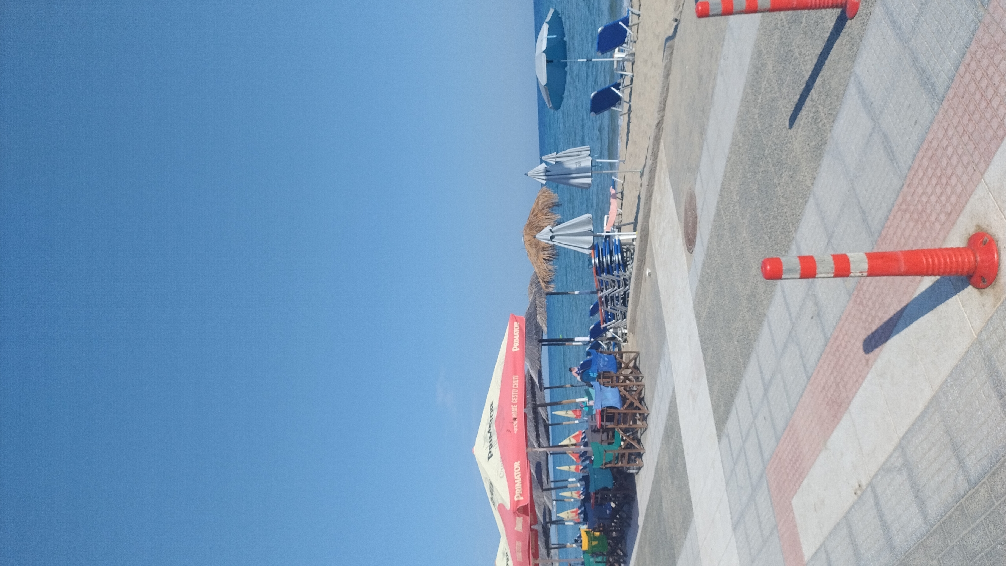 The image depicts a sunny day at a beachside establishment in Perea, Greece. In the foreground, a pedestrian walkway paved with patterned tiles leads towards the beach. Two red and white striped bollards stand on the walkway, casting shadows.
The middle ground features a beach with numerous lounge chairs and umbrellas. A section of what appears to be an outdoor dining area is visible, with tables and chairs set up under a large red and white umbrella bearing the "PRIMATOR" logo. Beyond the dining area, the sandy beach stretches towards the sea. Several blue lounge chairs and umbrellas are arranged on the sand.
The background is dominated by the calm, blue sea and a clear, bright blue sky. The overall scene conveys a relaxed atmosphere of a seaside resort.