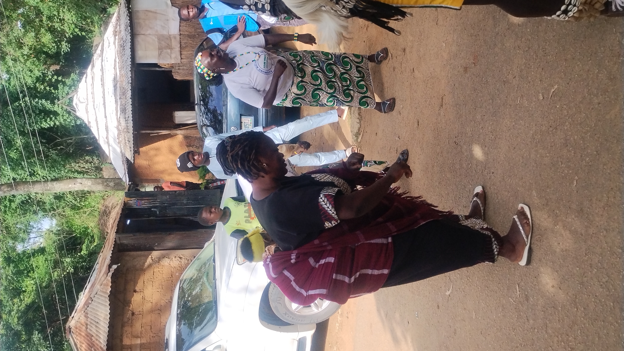 A group of people are gathered outdoors in a village setting, possibly in Jos, Nigeria. In the foreground, a woman with dreadlocks is bent over, her hands near the ground. She is wearing a dark shirt, a patterned skirt, and a maroon wrap around her shoulders and back. Two people are standing behind her, and to the right, a woman in a white shirt and a green and white patterned skirt is looking to her left and smiling. Behind them, several buildings with thatched and corrugated iron roofs are visible, along with a white SUV and a dark car. The ground is a dirt path, and lush green trees are in the background.