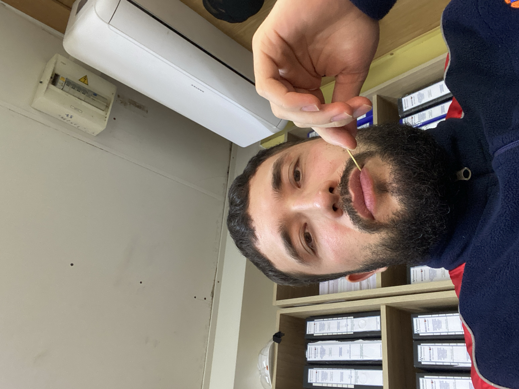 A man with a dark beard and short dark hair is in the foreground, holding a toothpick to his lips. He is wearing a dark blue fleece jacket with red trim. Behind him, shelves are filled with binders. A white air conditioning unit is mounted on the ceiling, and a white electrical box is to its left. The scene appears to be an indoor office or workspace. The image is a selfie, taken from a low angle, looking up at the man. There are a few small dark spots on the ceiling. The lighting is natural, coming from overhead.