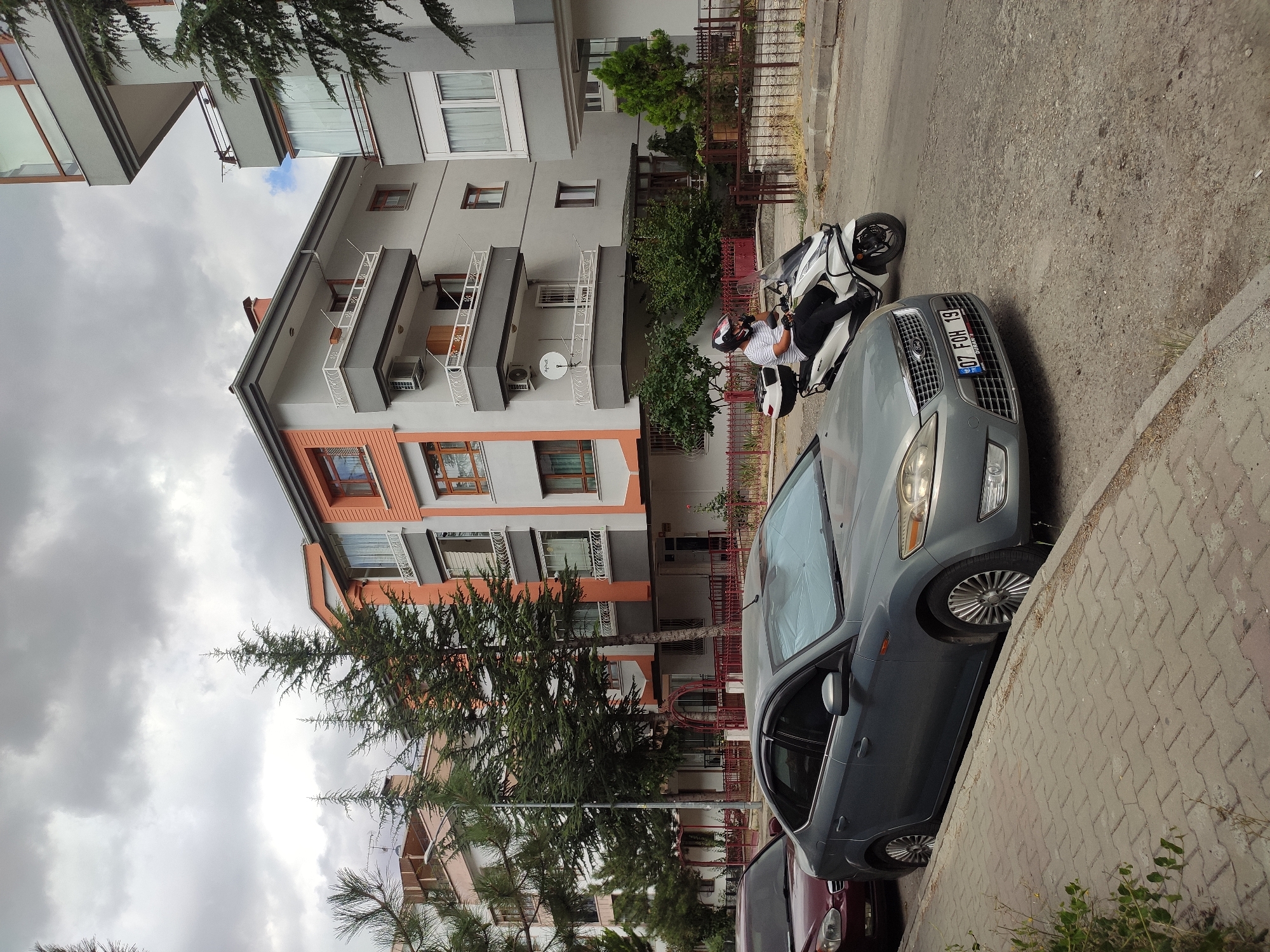 In the city of Mamak, Turkey, on a cloudy day, a street scene unfolds with apartment buildings and parked vehicles. A gray Ford sedan is parked on the left side of the road, facing right. To its right, a white scooter is being ridden by a person wearing a white and black striped shirt, black pants, and a helmet. The rider is in motion, with their left hand on the handlebars and their right hand near their face. Behind the scooter and sedan, a modern apartment building with orange and gray facades and multiple balconies stands. In the background, more apartment buildings and tall evergreen trees line the street. The ground is paved with interlocking concrete blocks along the sidewalk and asphalt on the road. A metal fence runs along the front of the apartment building.