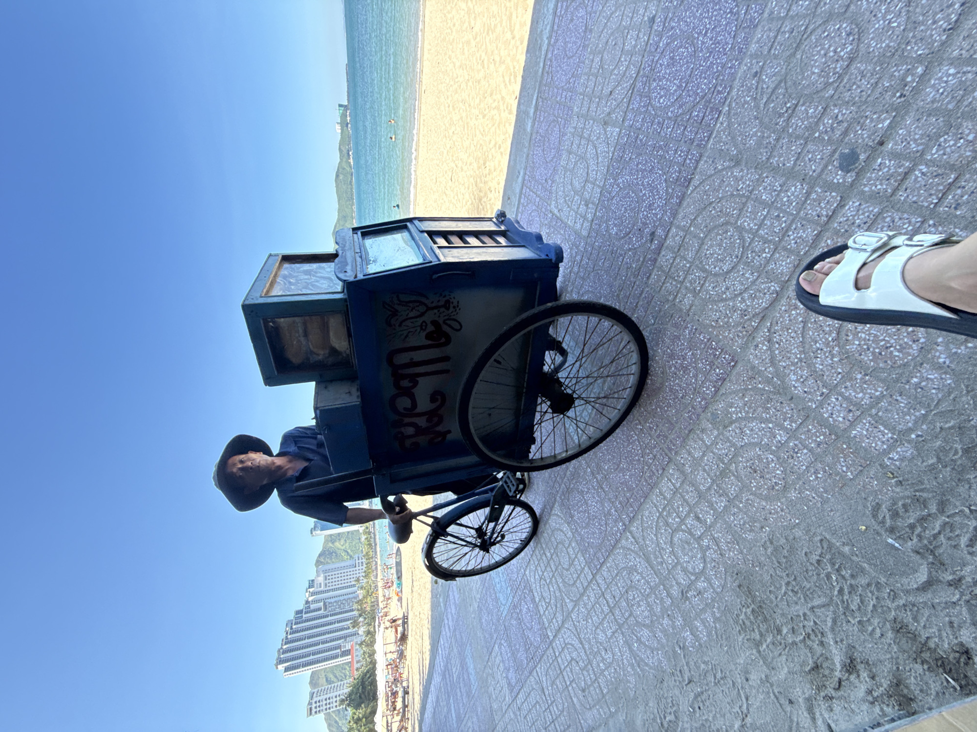 The image shows a man wearing a hat and a blue shirt, standing next to a blue bicycle cart on a paved walkway. The cart has a sign with decorative lettering that reads "Ream". In the background, there is a sandy beach with people and beach umbrellas, and beyond that, the ocean and distant green hills with buildings. The sky is clear and blue. The foreground shows a person's foot wearing a white sandal. This scene appears to be on the coast in Bac Nha Trang Ward, Vietnam, likely near the beach area.