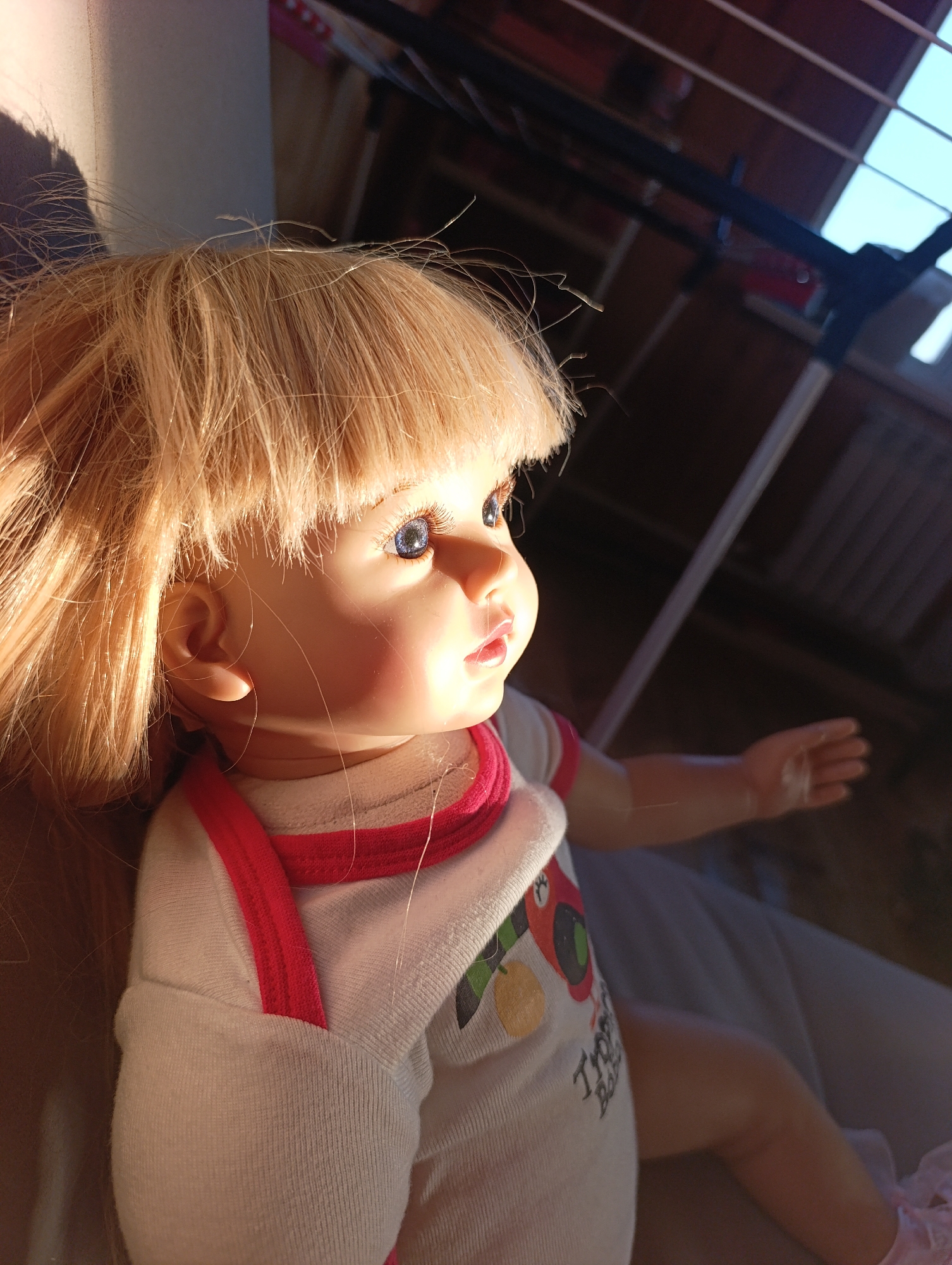 A plastic doll with blonde hair and blue eyes is sitting on a white surface, likely a bed. The doll is wearing a white shirt with red trim and a colorful design. The background is a room with a window and some furniture or equipment overhead. It is difficult to tell the precise location within Krasnoyarsk, Russia, without further information or context.