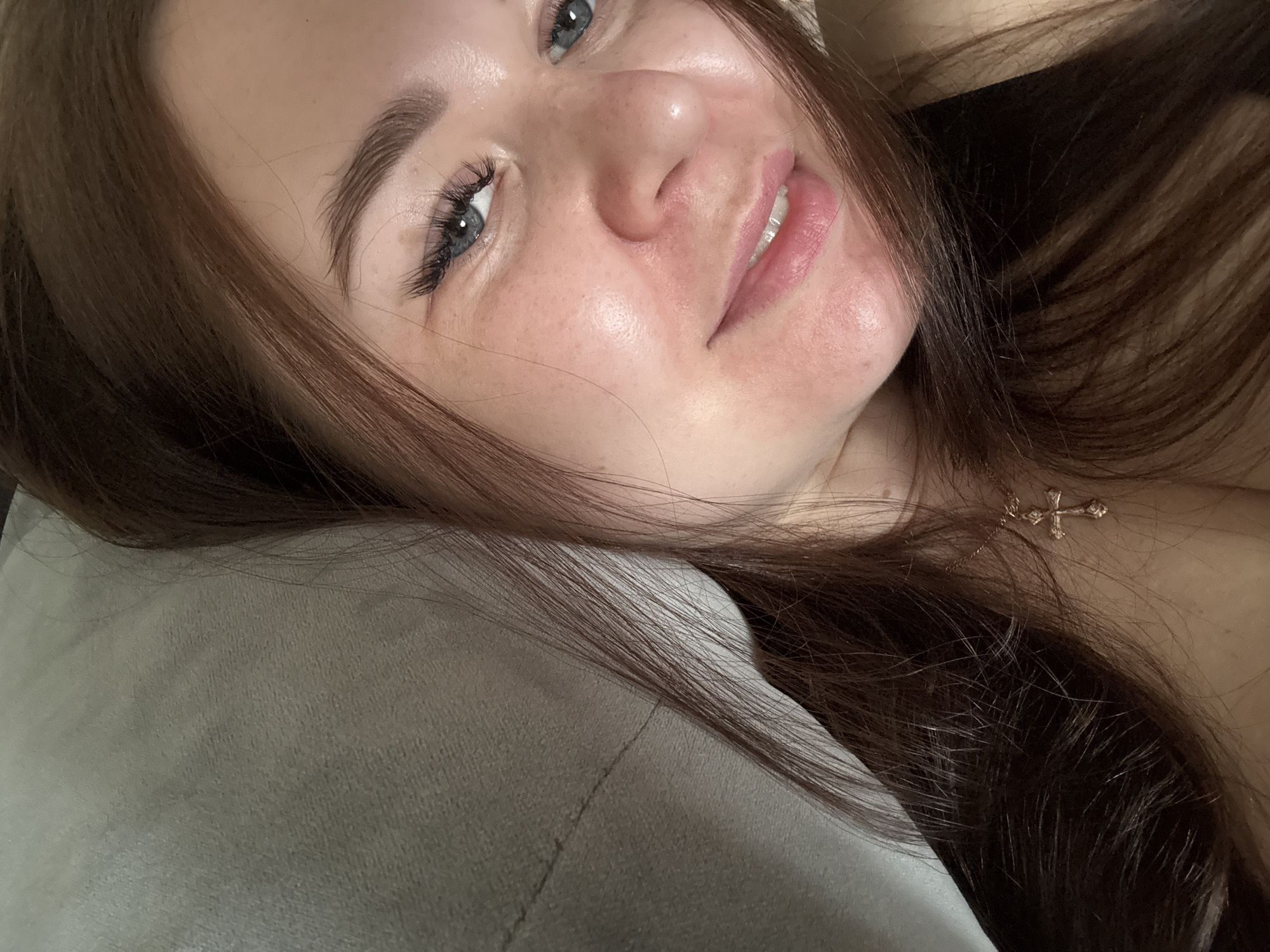 This is a close-up, selfie-style photo of a young woman with long, dark brown hair, lying on her side. She is wearing a delicate gold necklace with a cross pendant. Her eyes are blue and her lips are a soft pink. The background appears to be a bed or pillow, with a light gray texture. The lighting is soft and natural. There is no identifiable information about the specific location within Chaikovsky, Russia.