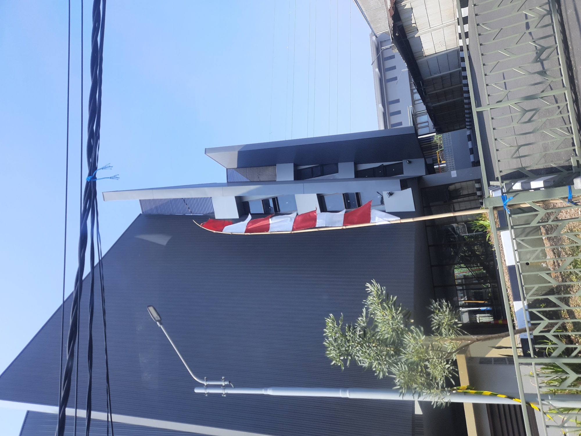 A bright, sunny outdoor scene in Bandung City, Indonesia, features a modern, dark gray, multi-story building with angled architectural elements. A tall, gray street lamp stands in the foreground to the left of the building, and a small, leafy green tree grows near its base. A flagpole with the Indonesian flag, displaying red and white horizontal stripes, is visible to the right of the street lamp. The building's facade has numerous windows. In front of the building, a metal fence runs horizontally across the lower portion of the image, with a paved area behind it. Overhead, several black power lines are strung across the clear blue sky. In the background, another lighter-colored building with a row of windows is visible, along with a smaller structure that appears to be a workshop or shed with a corrugated roof.