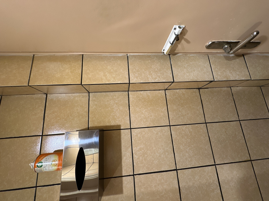 The image displays a ground-level view of a tiled floor meeting a plain wall, likely in a restroom or public facility in Nea Erithrea, Greece. The floor is covered with light brown or beige square tiles featuring a mottled texture and dark grout lines. The wall above the tiles is a uniform, light tan color.
Several objects are mounted on the wall. Towards the upper right, a silver-toned metal fixture, possibly a grab bar or a door stop mechanism, extends with a lever or handle. To its left, a white bracket with a dark clip or hook is also affixed to the wall.
In the bottom left section of the frame, a rectangular, reflective silver dispenser rests on the floor, positioned against the wall. It has a distinctive oval-shaped opening on its front. Partially visible inside this dispenser is an orange and white spray can, which appears to be an air freshener or disinfectant. The word "Pure" is discernible on the visible part of the can's label.
The scene is static, with no people or active events taking place. The lighting is artificial, casting subtle shadows, particularly from the dispenser onto the tiled floor, consistent with an indoor, utilitarian environment.