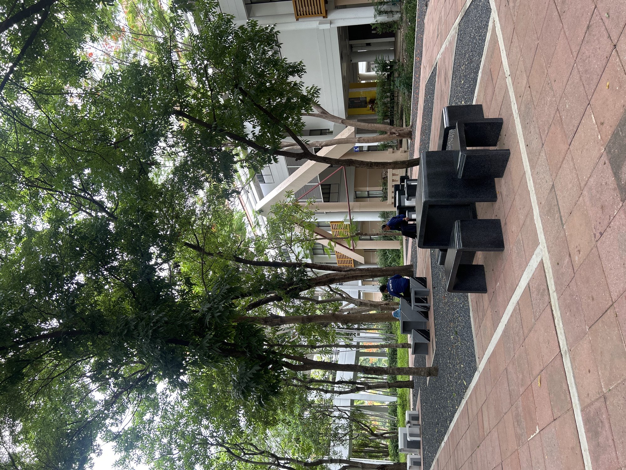 A wide shot captures an outdoor seating area with several modern, dark stone tables and benches. The area is paved with red tiles and surrounded by trees and gravel. In the background, a multi-story building with a white and beige exterior and angular architectural features is visible. Two individuals are seated at one of the tables on the right side of the frame. The person on the left is wearing a dark blue shirt, and the person on the right is also in a dark blue shirt, facing away from the camera. The lighting suggests an overcast day, with diffused light filtering through the trees. The scene appears to be a public space within a university campus or a similar institutional setting in Tha Khlong Town Municipality, Thailand.