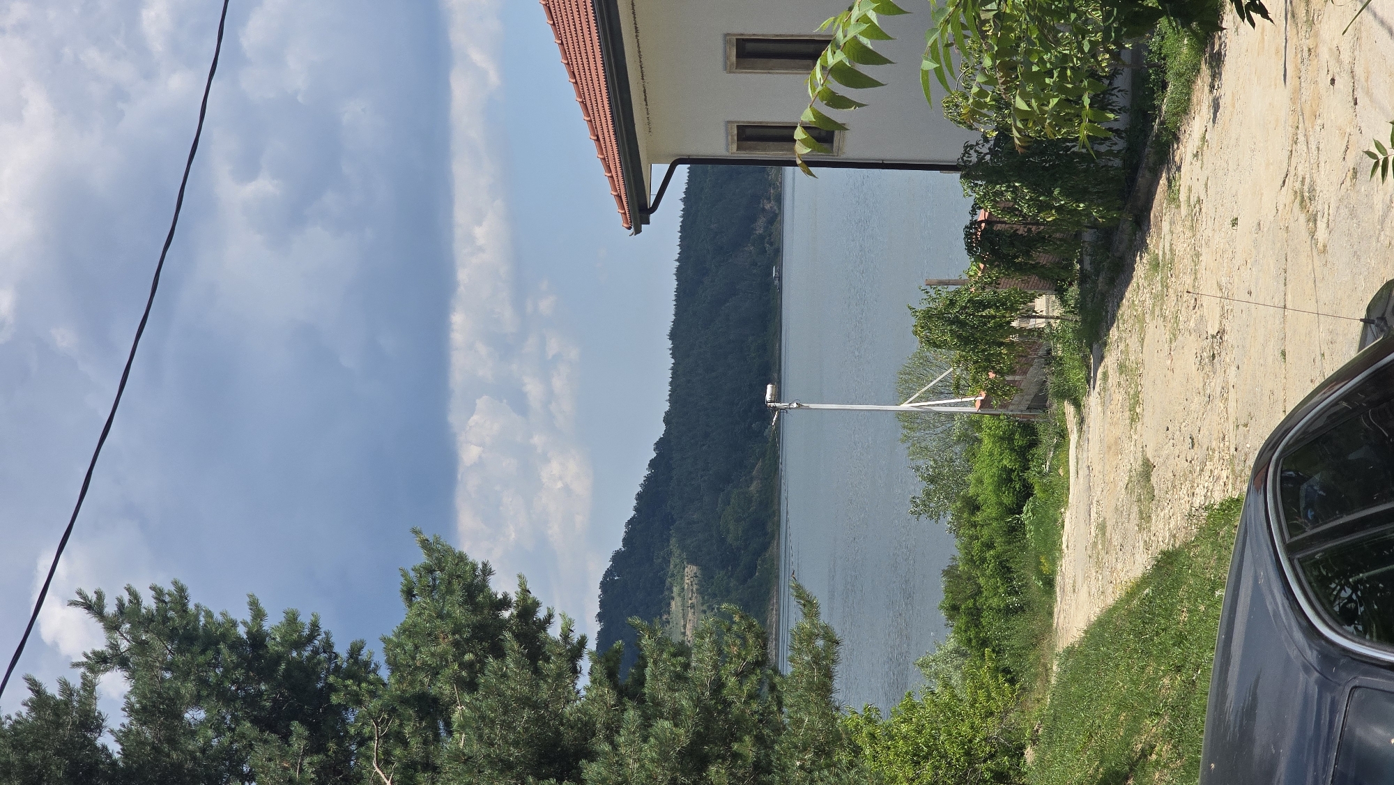 The image shows a view towards a body of water, likely the Danube River, from a property in Asparuhovo, Bulgaria. A dark-colored car is partially visible in the foreground on the left, with its roof and passenger side window showing reflections. A dirt and gravel path leads away from the viewer towards the water. On the right side of the path is the side of a white building with a red tile roof. Two windows are visible on the side of the building. Behind the building, a brick wall and green trees are present.
Beyond the path and the building, a large expanse of water stretches into the distance. Across the water, a densely forested hill or mountain range forms the horizon. The sky above is partly cloudy with a mix of dark and light gray clouds, indicating a somewhat overcast day. A single power line is visible diagonally across the upper portion of the image. A tall, white pole with a light fixture is positioned near the water's edge. Green trees and vegetation line the banks of the water and surround the path.