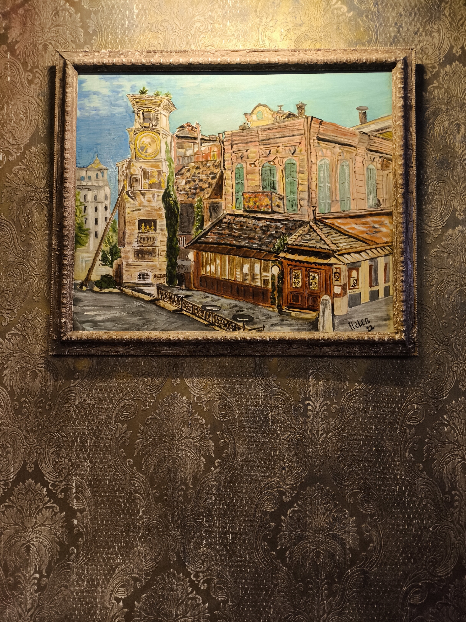 A framed oil painting hangs on a brown-patterned wall. The painting depicts a city scene, including a clock tower, buildings with shutters, and what appears to be a covered outdoor cafe. A signature "Helen" is visible in the lower right corner of the painting. The city appears to be Moscow, Russia, given the style of architecture. No people are visible in the image.
