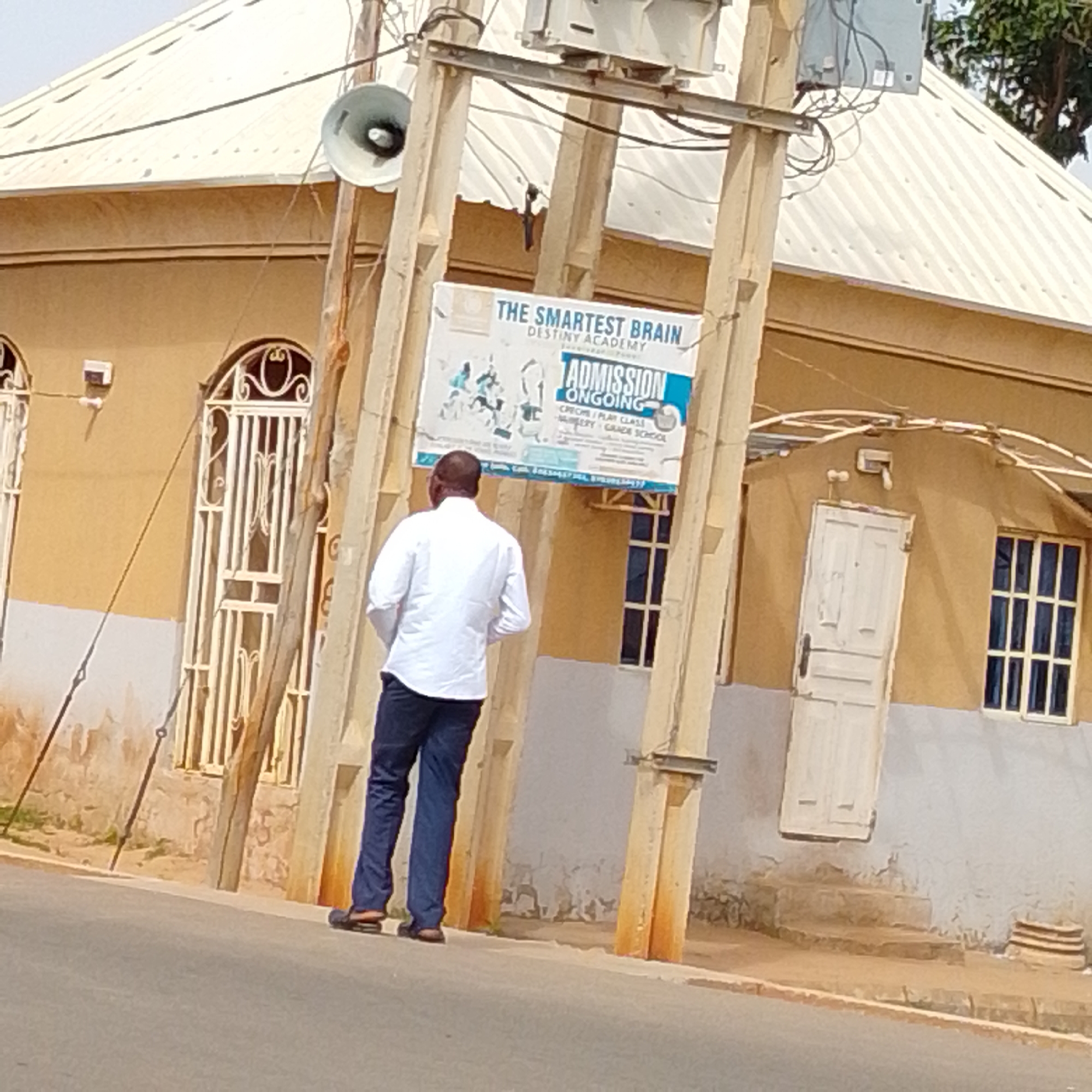 The image depicts a street scene in Gombe, Nigeria. A man wearing a white shirt and dark trousers stands facing away from the camera, looking at a sign. The sign, attached to a utility pole, advertises "THE SMARTEST BRAIN DESTINY ACADEMY" and states "ADMISSION ONGOING" for "CRECHE / PLAY CLASS / NURSERY / GRADE SCHOOL." The building behind the sign has a light brown facade with grey trim and barred windows. A white door with a step leading up to it is visible on the right side of the building. A metallic roof covers the building. Utility poles with wires and a speaker are also prominent in the foreground. The ground is paved with asphalt and a curb is visible. The overall environment appears to be an urban setting with a slightly weathered building and street infrastructure.