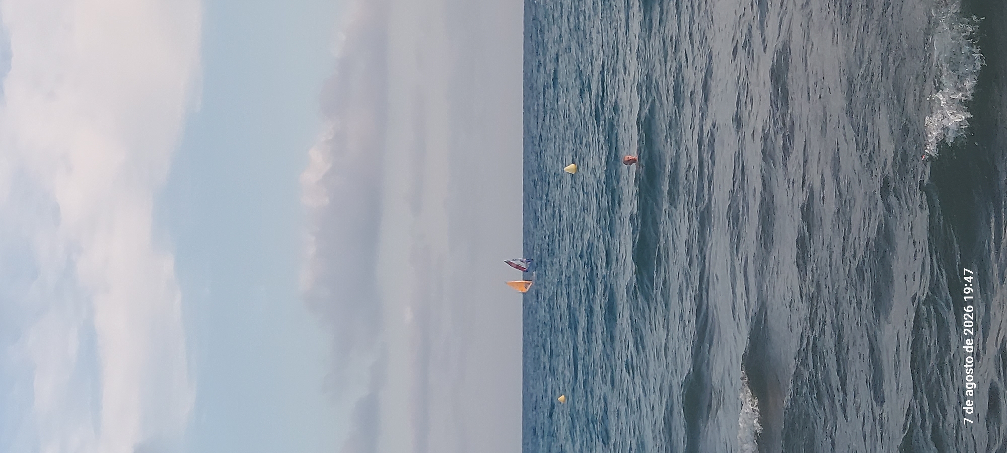 This image captures a view of the sea off the coast of Castelldefels, Spain, on August 7, 2026, at 19:47. The scene is dominated by the sea and sky. The sea is a deep blue, with visible waves and foam along the shore in the foreground. Two windsurfers, identifiable by their colorful sails (one orange, one red and blue), are visible in the middle ground of the sea. In the right middle ground, a person's head is just visible above the water, suggesting they are swimming. Two yellow buoys are also present in the water. The sky above is a light blue, with scattered white clouds that appear soft and diffused. The overall atmosphere is one of a calm, late afternoon at the beach.