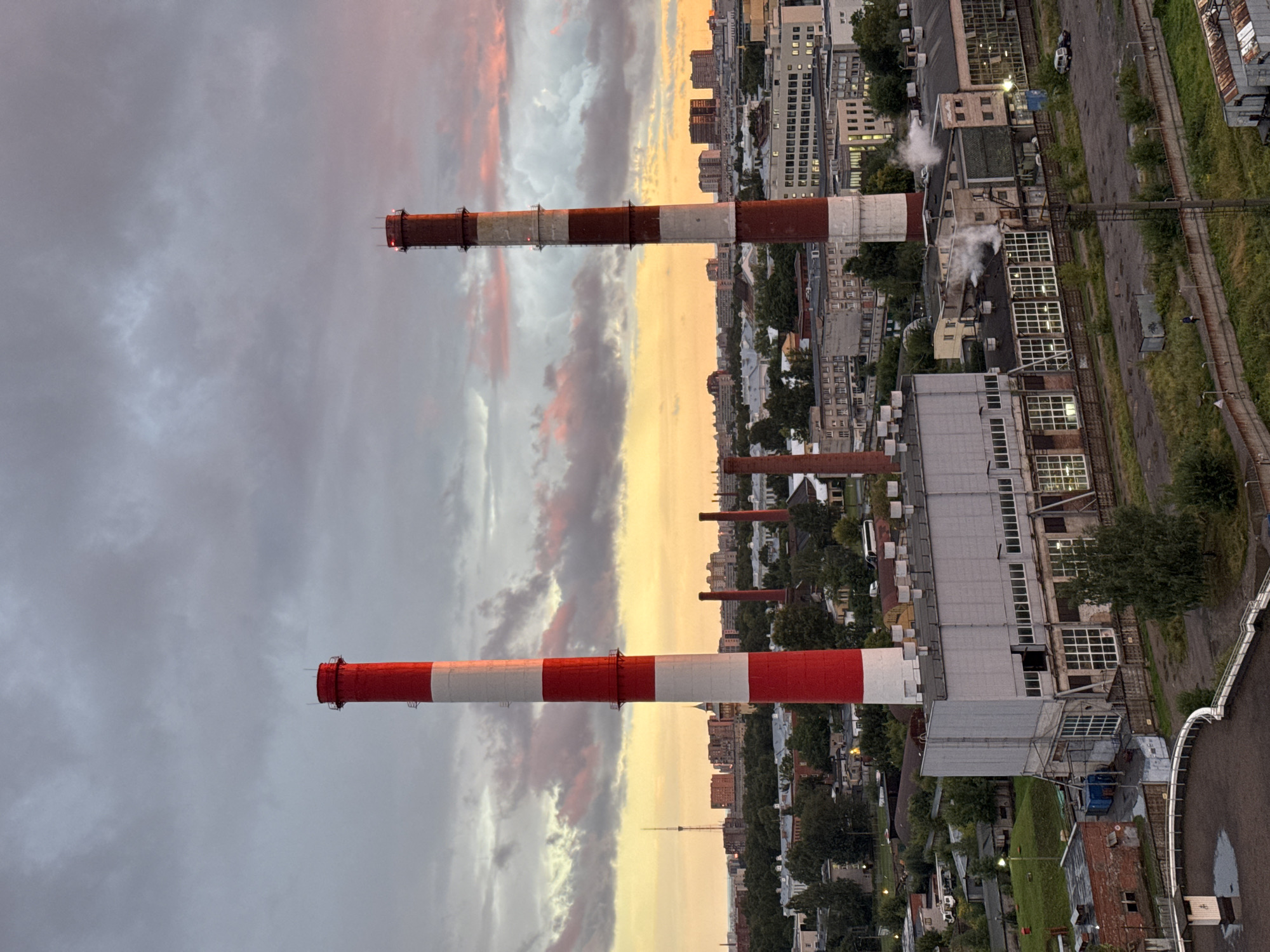 The image depicts an industrial landscape in Sinyavino, Russia, under a dramatic sky with colorful clouds illuminated by sunset. Dominating the foreground are two tall, red and white striped smokestacks, characteristic of a power plant or industrial facility. Below and surrounding the smokestacks are various buildings, including a large, industrial complex with large windows and a lighter-colored roof, and several smaller, older brick structures. Smoke or steam emanates from one of the buildings, suggesting ongoing industrial activity. In the distance, a cityscape with numerous buildings of varying heights is visible, along with a prominent telecommunications tower. Green trees and vegetation are interspersed throughout the scene, indicating an urban environment with some natural elements. A few vehicles, including cars and a bus, are present in the industrial area. The overall impression is one of a working industrial zone against a backdrop of a vibrant, natural sky.