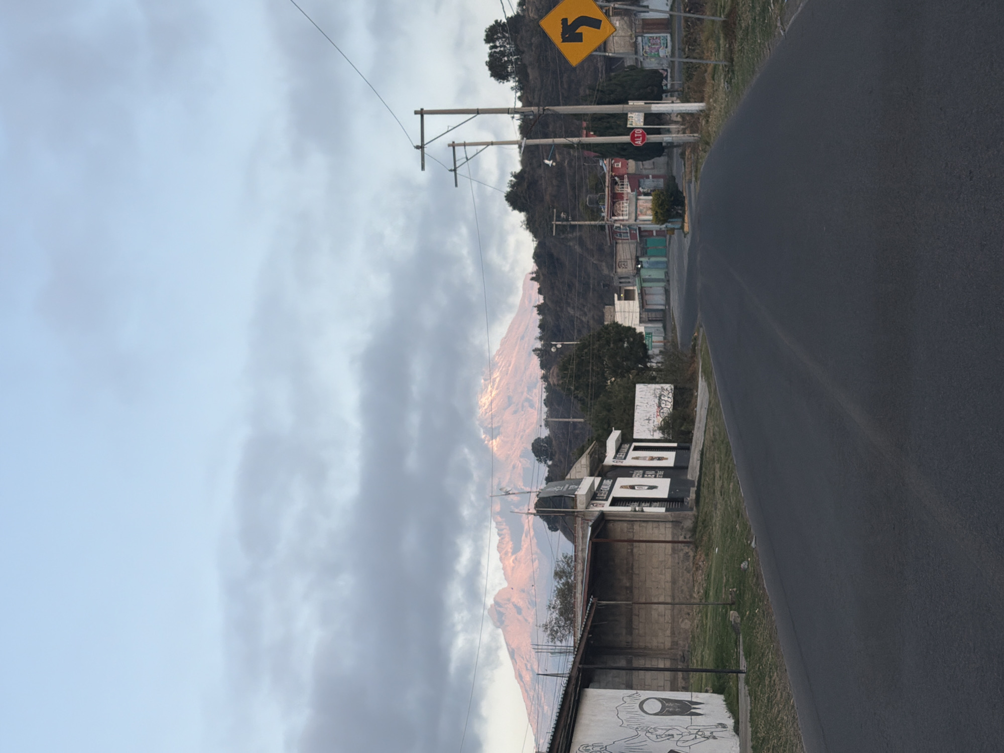 The image captures a street scene in San Pedro Yancuitlalpan, Mexico, featuring a view of a mountain range in the distance, likely illuminated by the rising or setting sun. The sky is partially overcast with grey clouds. A paved road curves through the foreground. Utility poles with power lines run along the roadside. A yellow road sign indicating a curve is visible. Buildings of varying heights and conditions line the street, and some have graffiti. A red "ALTO" stop sign is also visible. A wall and some vegetation is located along the side of the road.