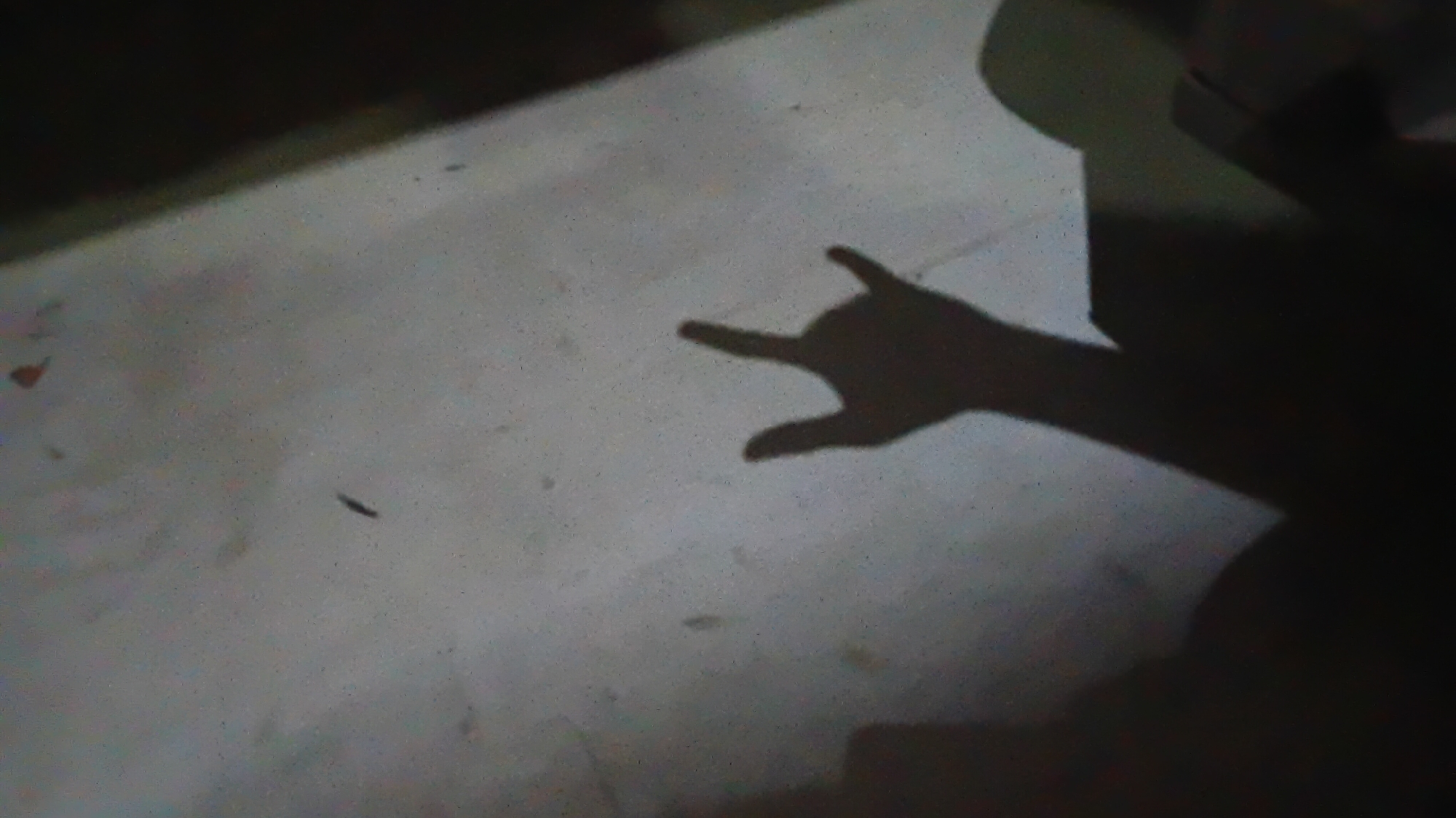 The image depicts a shadow of a hand performing the "rock on" gesture, with the index finger and pinky finger extended, against a textured, light-colored surface. The surface appears to be a wall or floor with a slightly gritty texture, possibly concrete or stucco, and exhibits some dust or small debris. To the left of the hand's shadow, a larger, more amorphous shadow is visible, suggesting the presence of a person or another object casting a shadow. To the right of the hand's shadow, another shadow is partially visible, possibly of another person or an object. The lighting suggests an indoor or sheltered environment with a light source positioned to create these shadows. There is no specific indication of the city of Prabumulih, Indonesia, within the image itself. The image quality is somewhat grainy, and some areas are underexposed, making it difficult to discern fine details.
