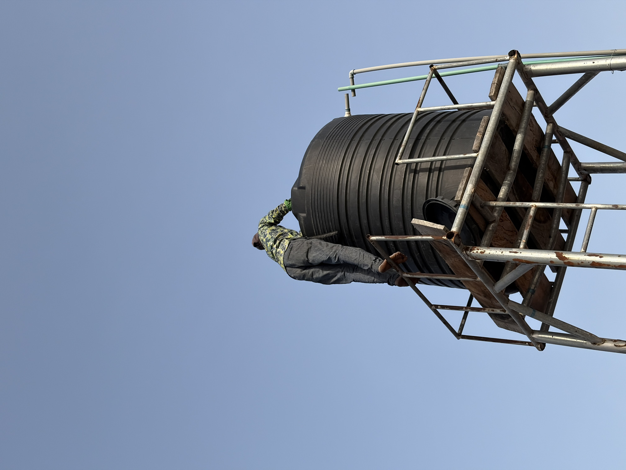 A low-angle shot shows a black plastic water tank supported by a metal frame. A person, wearing a patterned long-sleeved shirt and pants, is positioned on top of the tank. The person's feet are visible, positioned on the frame. The background is a clear, light blue sky. The location is Igboefon, Nigeria.