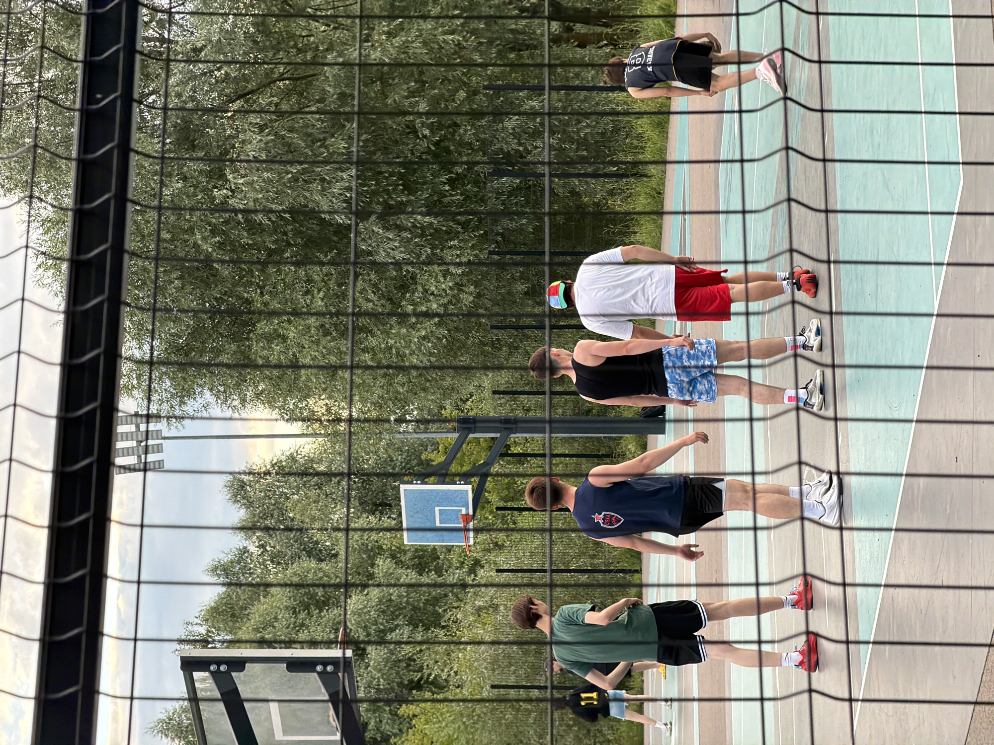 The image shows a group of young men playing basketball on an outdoor court. The court has a light blue and green surface with white lines. A net is in the foreground, partially obscuring the view of the players. Trees and foliage surround the court, and a basketball hoop is visible in the background. The time of day appears to be late afternoon or early evening, with soft lighting. The weather seems pleasant, as the players are dressed in athletic wear. The atmosphere appears casual and recreational. The people are all wearing athletic shorts and t-shirts or jerseys. One player is wearing a dark blue jersey with a red and white logo. Another player is wearing a white t-shirt and red shorts. A third player is wearing a black tank top and blue and white patterned shorts. A fourth player is wearing a green t-shirt and black shorts. A fifth player in the background is wearing a black jersey with white lettering and a number. The location is an outdoor basketball court in Kazan, Russia.