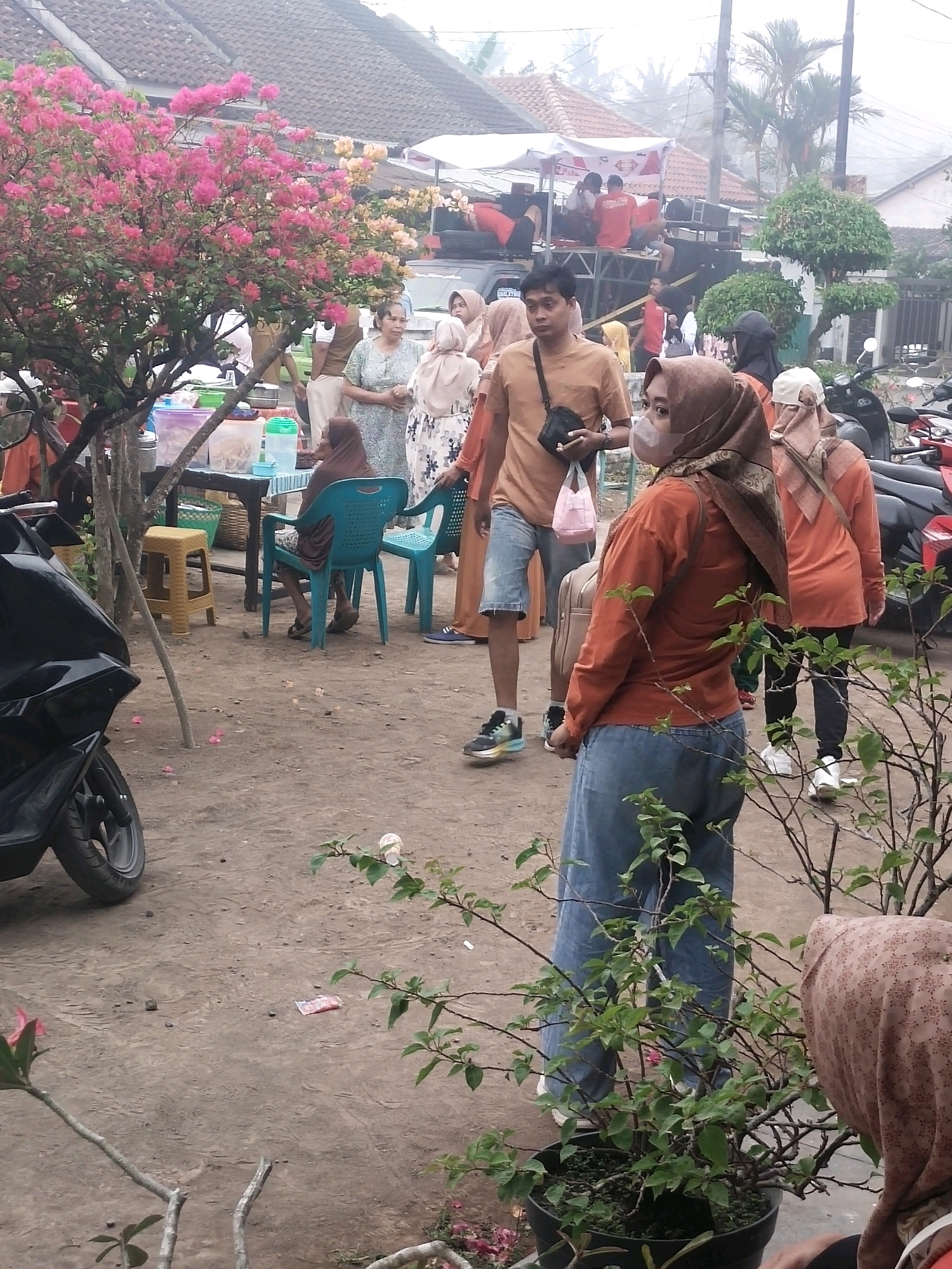 The image depicts an outdoor scene in Ambulu, Indonesia, likely a community gathering or market. In the foreground, a person wearing a rust-colored long-sleeved shirt and blue jeans stands facing away from the camera, with a brown backpack slung over their shoulder. To their right, another person in a similar rust-colored shirt and a headscarf is visible from the back. In the background, a group of people are gathered around a stage or platform adorned with speakers and banners, suggesting a performance or event is underway. A vibrant pink bougainvillea bush is prominently featured on the left side of the image, adding a splash of color to the scene. The ground is dirt and gravel, with several parked motorcycles and a black scooter visible. Tables with various items, possibly food or merchandise, are set up, with plastic containers and baskets present. The overall atmosphere appears casual and lively, with a sense of community engagement.
