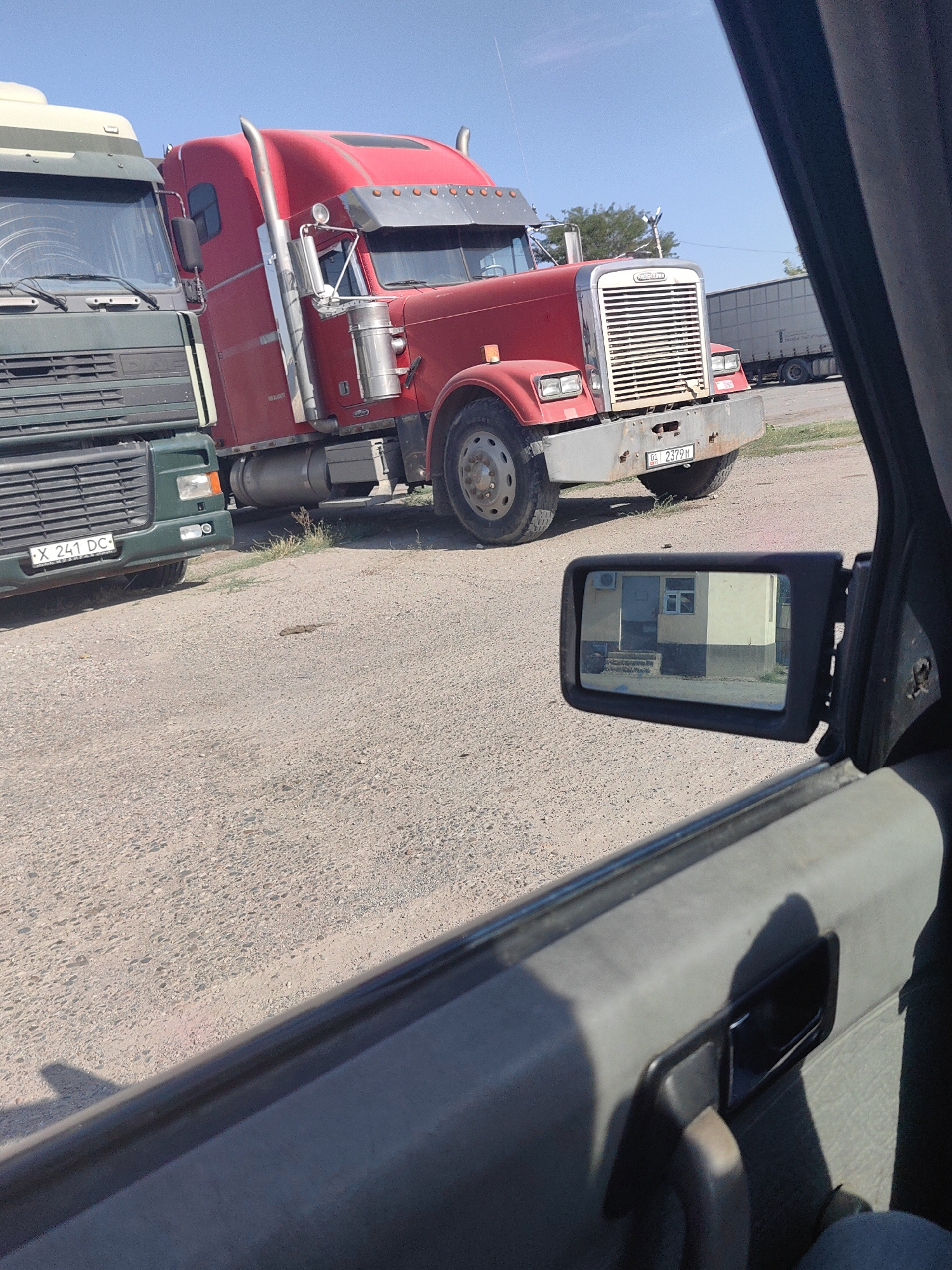 The image shows two large trucks parked on a gravel lot. The truck in the foreground is red and appears to be a Freightliner model. It has a large chrome grille and a license plate that reads "01 2379 M". To the left of the red truck is a green truck, possibly a DAF model, with a license plate that reads "X 241 DC". The scene appears to be outdoors in a sunny environment, with a clear blue sky. The image is taken from inside a vehicle, as evidenced by the car door and side mirror visible in the foreground. The side mirror reflects a building with a light-colored facade, a doorway, and a window. The presence of the license plate format "01" suggests the location might be the Turkistan Region of Kazakhstan, which includes the city of Shymkent. The overall impression is that of a truck stop or a parking area for commercial vehicles in Kazakhstan.