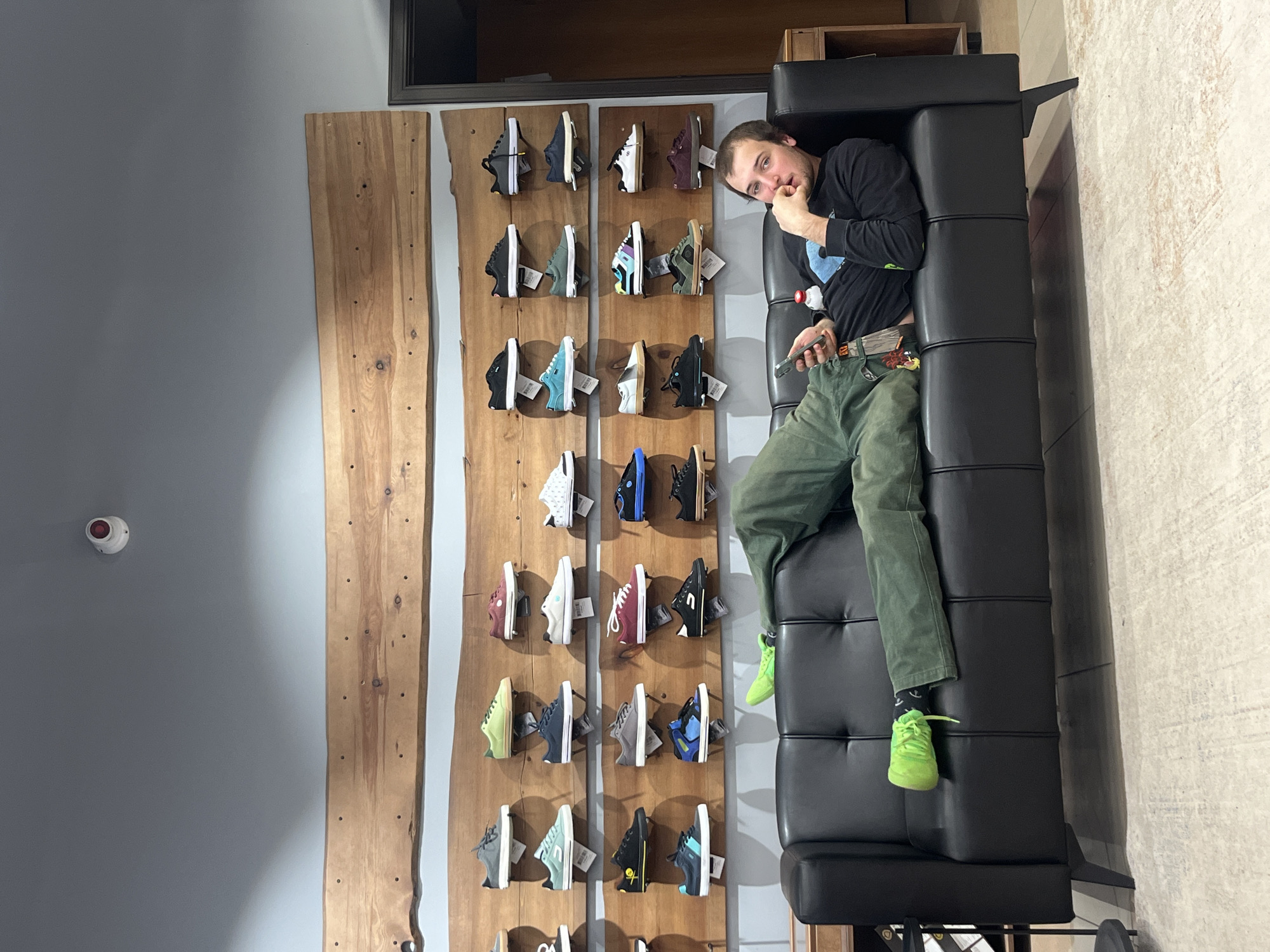 A man is reclining on a black leather couch next to a wall display of shoes in a retail setting. He is wearing a black long-sleeved shirt, green pants, and neon green shoes. He is holding a mobile phone and has his hand near his mouth as if thinking or eating. The shoe display consists of rows of sneakers mounted on wooden panels, affixed to the wall. The environment seems to be a shoe store or a store with a curated sneaker collection. There is a security or smoke detector on the wall. The location is most likely not Krasnoyarsk, Russia, as the image shows a retail environment more common in Western countries. There is no readily apparent evidence to indicate the city of Krasnoyarsk, Russia.