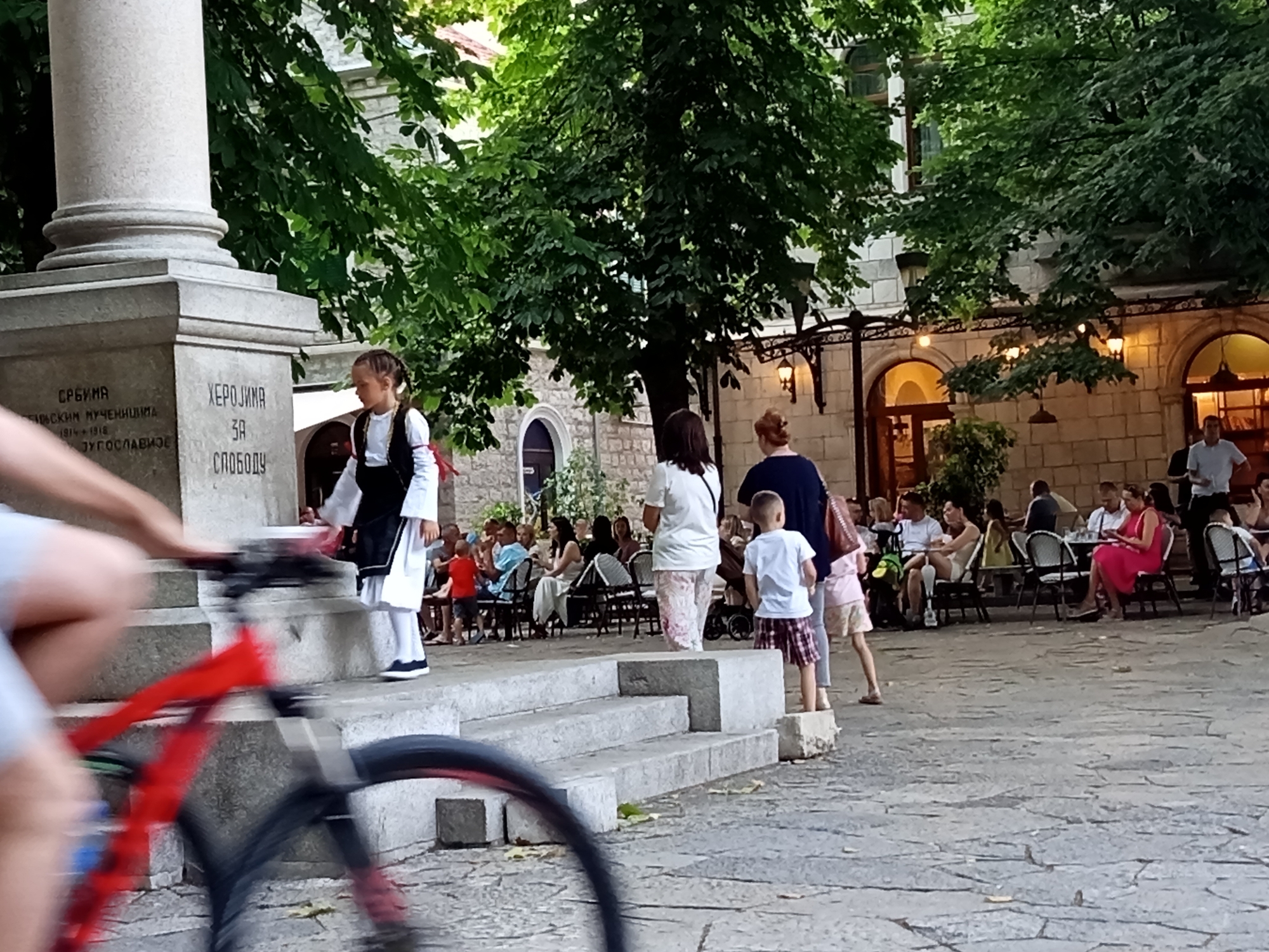 In Trebinje, Bosnia and Herzegovina, at the monument dedicated to Herzegovinian heroes, a young girl in traditional attire stands near the monument's inscription. A bicycle, with its front wheel out of focus due to motion blur, is in the foreground. In the background, people are seated at tables outside a building, suggesting a cafe or restaurant. The setting is an outdoor public space with trees providing shade.