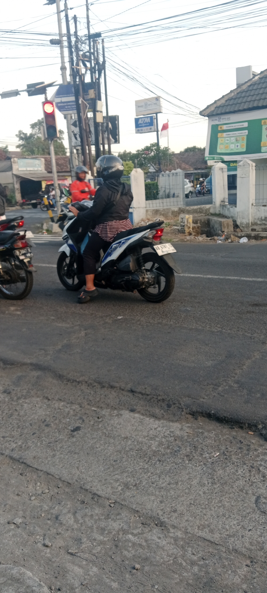The image shows a street scene in Maguwoharjo, Indonesia, under a bright, overcast sky. In the foreground, a person wearing a black jacket and a black helmet is riding a white and blue motorcycle. The rider is wearing striped pants and dark sandals. The motorcycle has a license plate that reads "AR 6781 YZ". To the left, another motorcycle is partially visible, with a rider wearing a dark helmet. In the background, there are utility poles with numerous wires, a traffic light displaying red, and signs for "BANK BPD DIY" and "ATM". A red and white Indonesian flag is also visible. Several buildings line the street, including one with a tiled roof. The road surface is asphalt, with some visible cracks and debris.