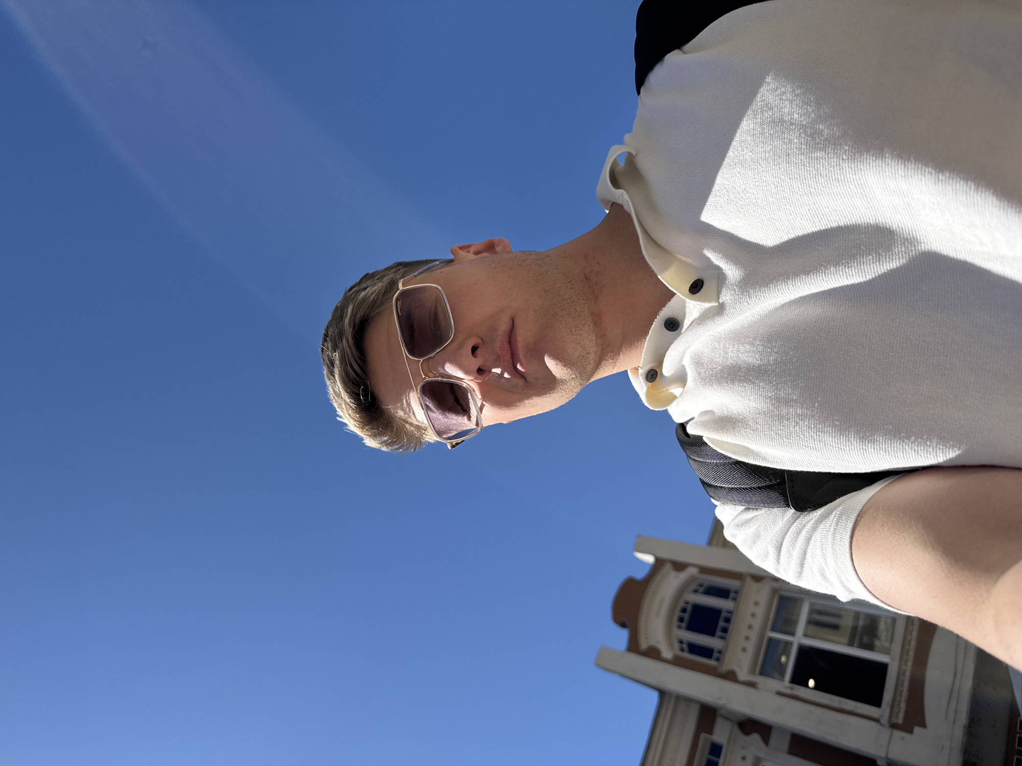 A low-angle selfie photograph shows a light-skinned person wearing sunglasses and a white shirt with black buttons. The person appears to be outdoors on a sunny day, with a clear blue sky filling much of the background. In the bottom right corner, a portion of a building with a distinctive window arch is visible. The building appears to be older and made of light-colored stone with dark trim. Based on the architectural style of the building in the bottom right corner of the image, it is likely located in Plovdiv, Bulgaria.
