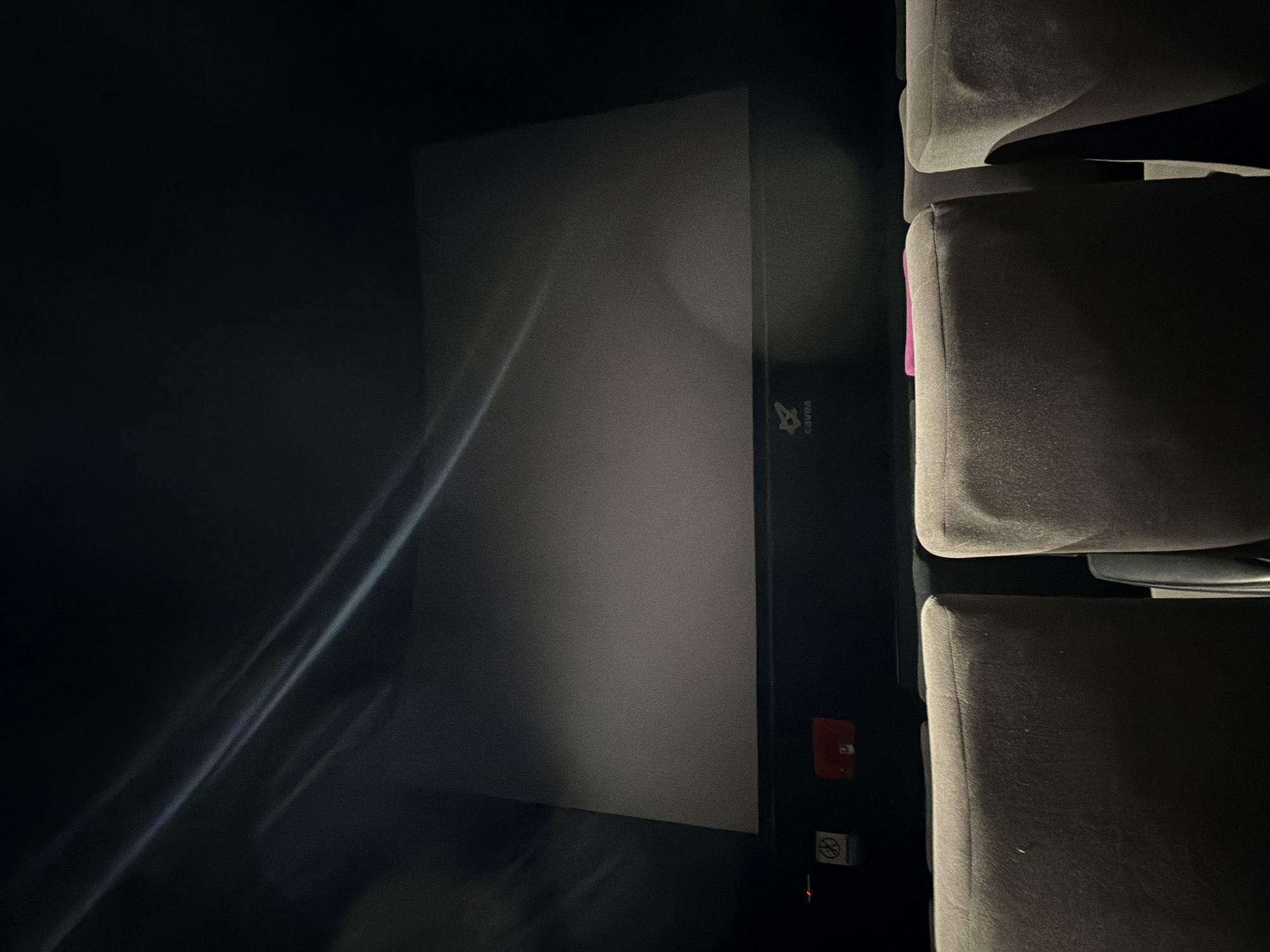 The image shows a cinema screen with the Cavea cinema logo visible, presumably located in Tbilisi, Georgia. The screen is blank, and the cinema is dark, with light reflecting off the screen surface. The composition includes cinema seats, positioned to the right of the frame. The room appears to be empty. A red light and a no-smoking sign are visible on the wall beside the screen.