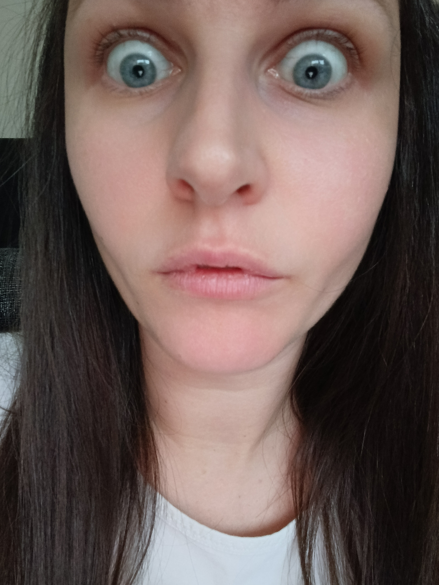 A close-up, slightly off-center, high-angle shot of a woman's face dominates the image. The woman's face is pale and without makeup. Her eyes are wide open, with large blue irises and pupils appearing dilated. She appears to be surprised. Her nose is slightly upturned, and her lips are slightly parted with a neutral expression. The woman has long, dark brown hair, with strands framing the sides of her face. She is wearing a white t-shirt. The location is Unknown, Unknown. The background is out of focus.