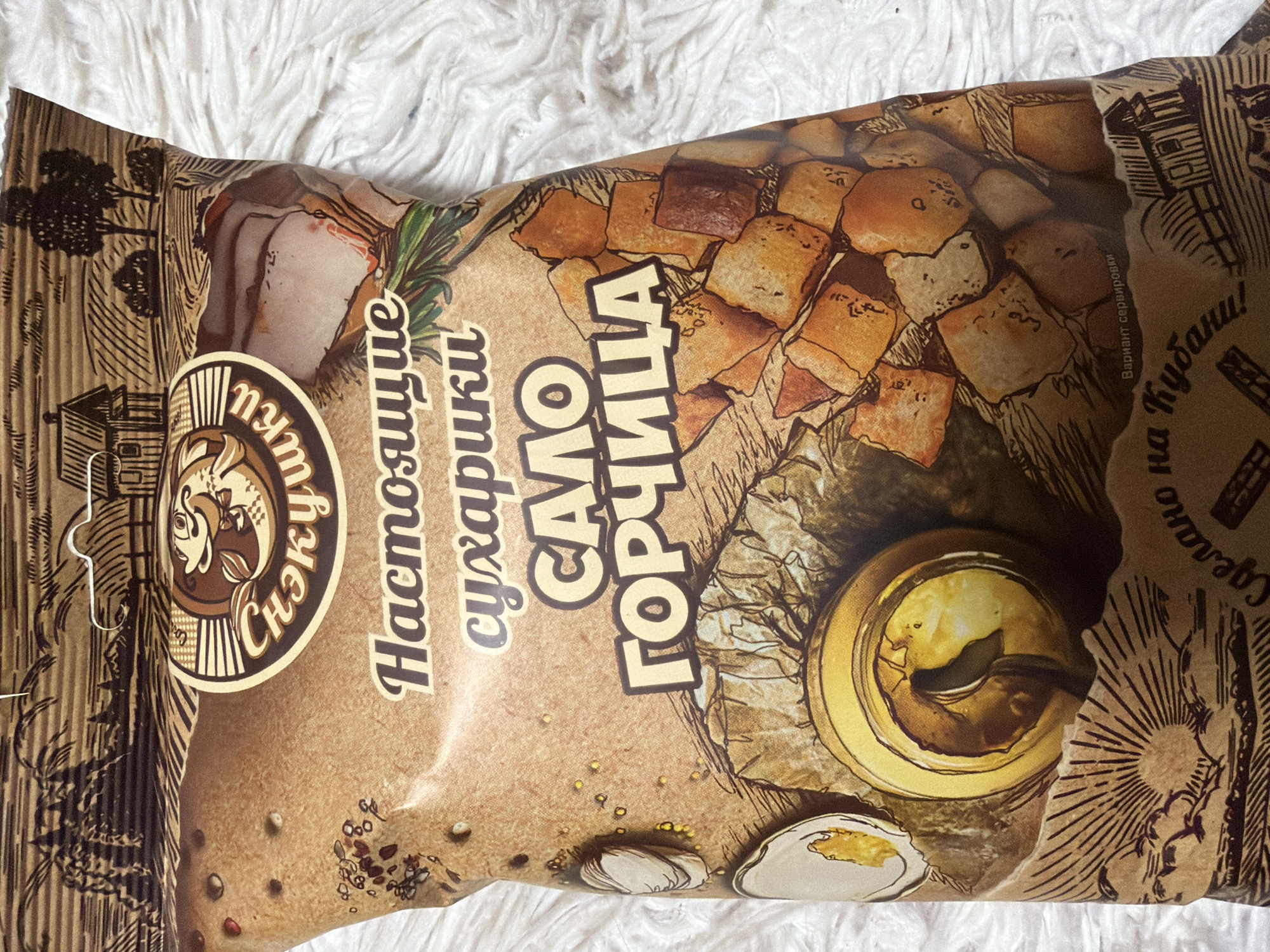 The image shows a package of "Snezhki" brand croutons. The flavor is "lard with mustard." The text on the packaging is in Russian. The text "Сделано на Кубани!" translates to "Made in Kuban!" There is no indication of Izhevsk, Russia in this image.